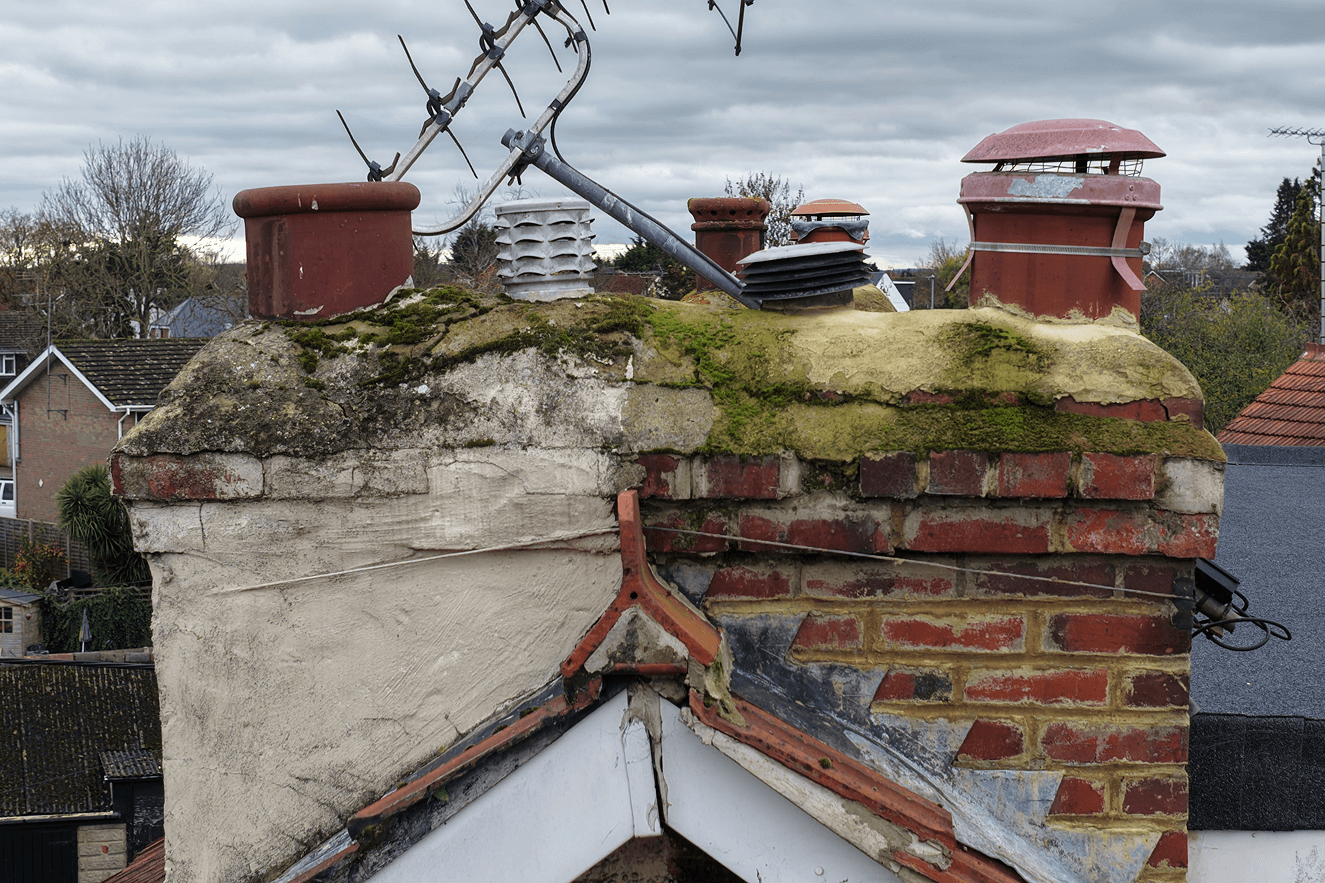 Close-up of a weathered chimney on a roof, with moss and signs of aging, surrounded by other rooftops and trees under a cloudy sky.