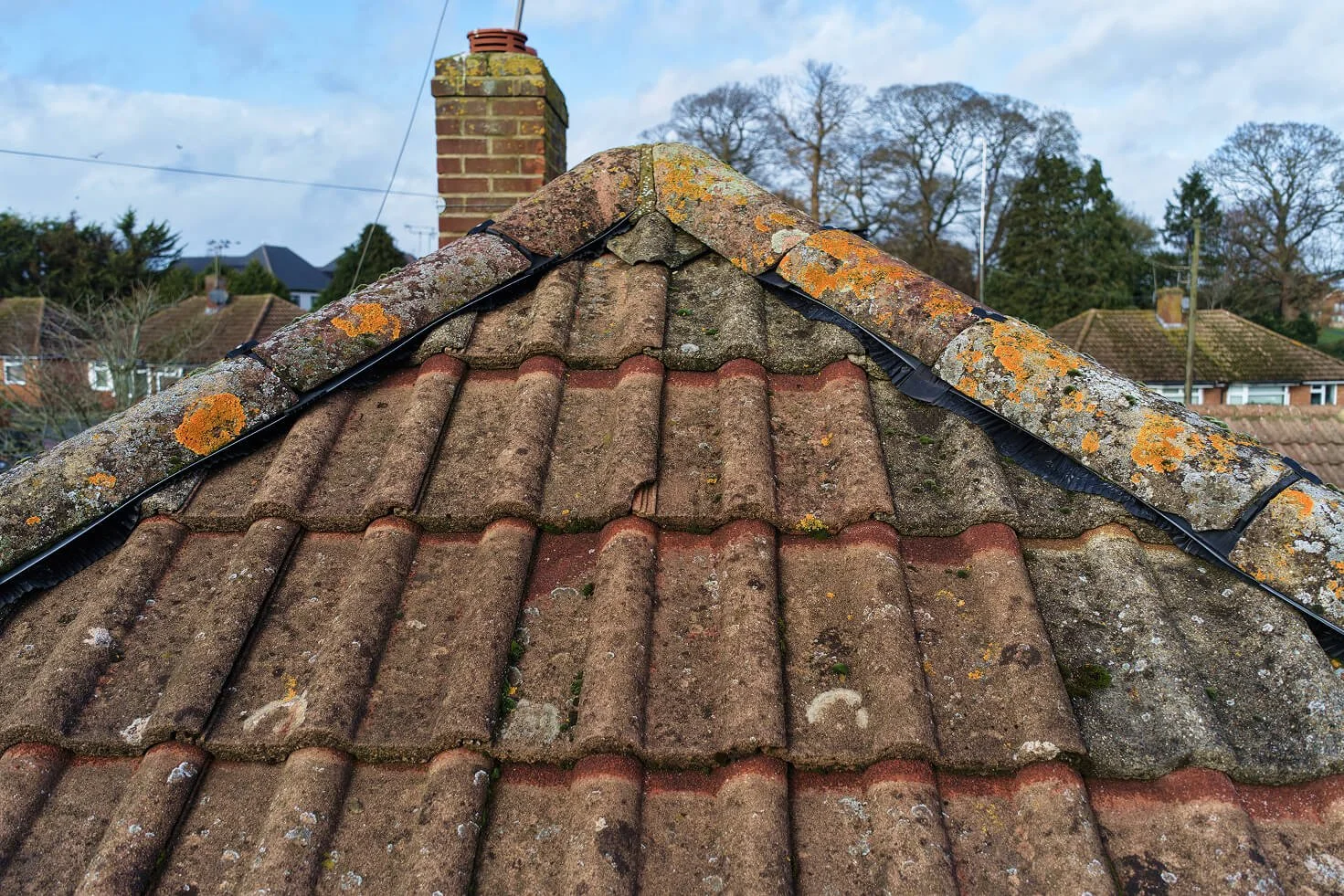 Close-up view of a moss-covered, weathered tiled roof with a chimney in the background, situated in a residential neighborhood with other houses and trees.