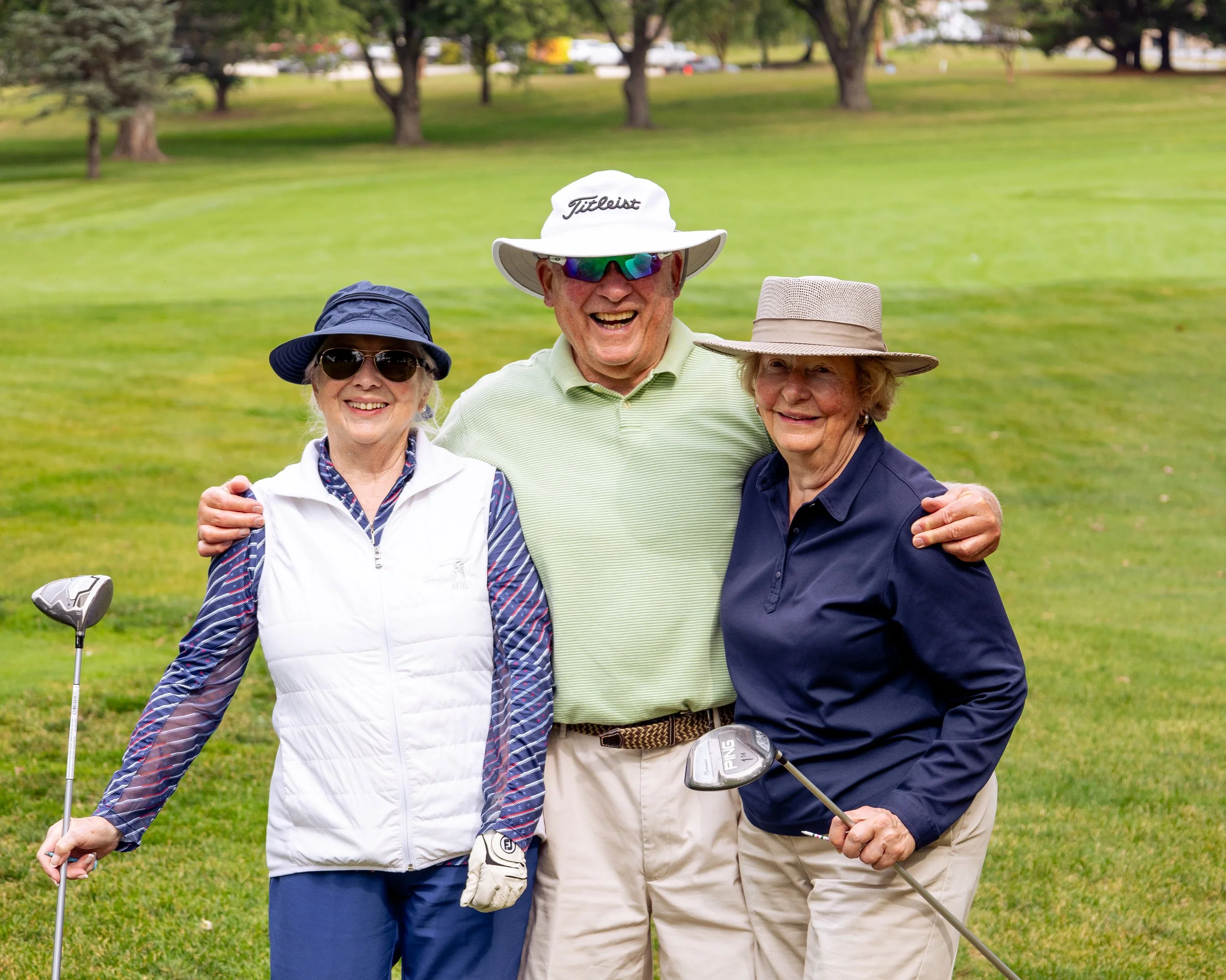 Three senior individuals standing on a golf course, smiling, holding golf clubs, with trees in the background.