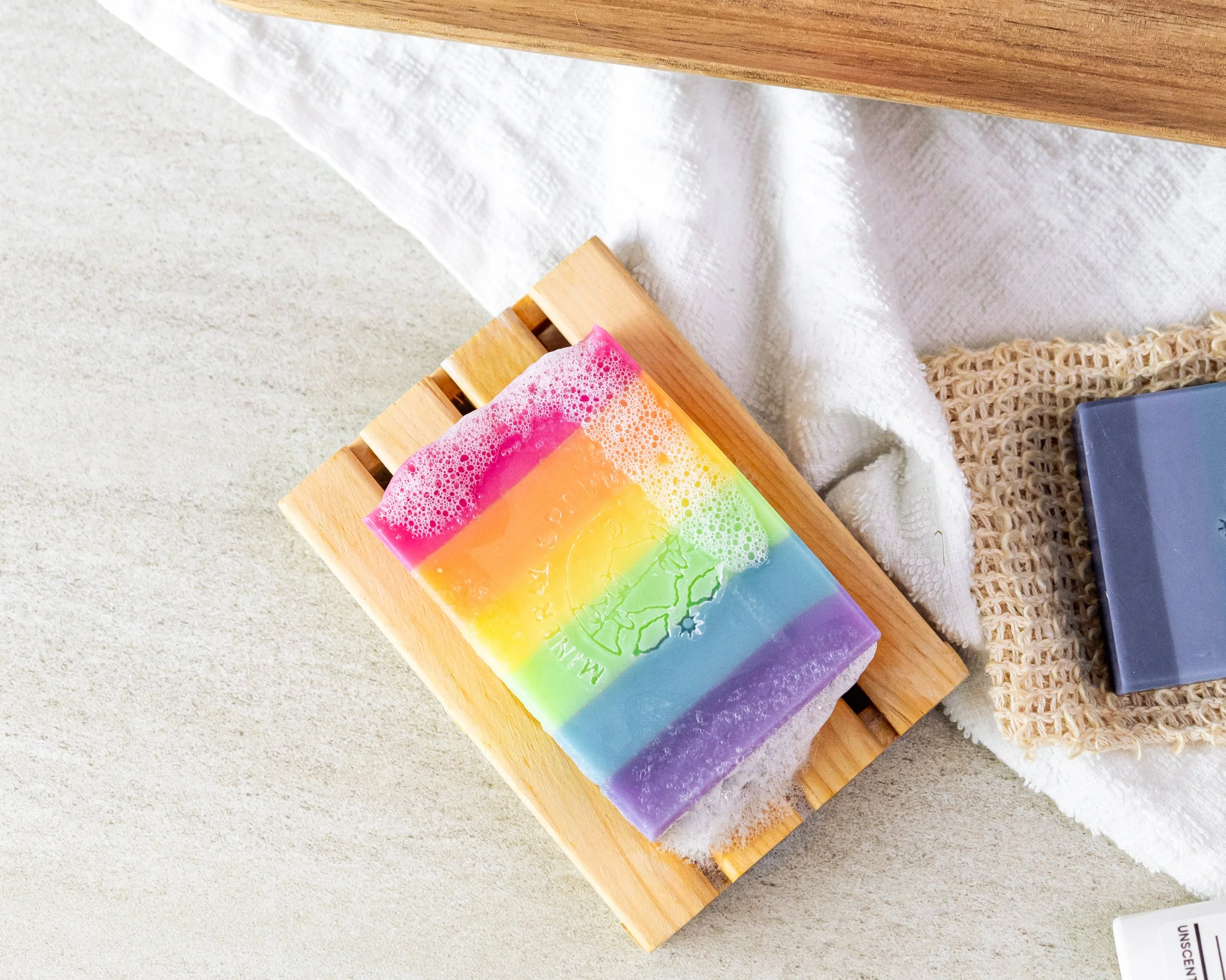 Colorful rainbow soap bar with a mountain logo and the words 'Make Mountains' on a wooden soap dish, surrounded by a white towel, a beige scrubby, a dark blue box, and a white surface.