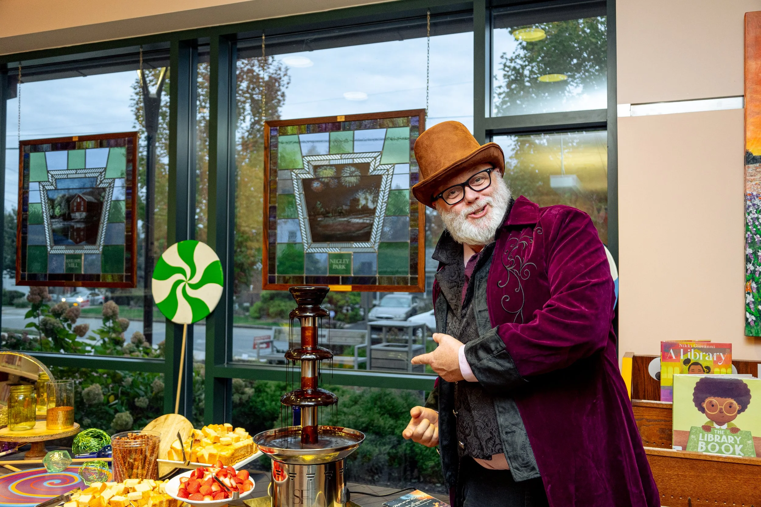 A man with a white beard, glasses, wearing a brown hat and a purple velvet coat, standing inside an art gallery or bookstore, pointing at a chocolate fountain on a table with cheese and strawberries, with stained glass artwork and books about libraries in the background.