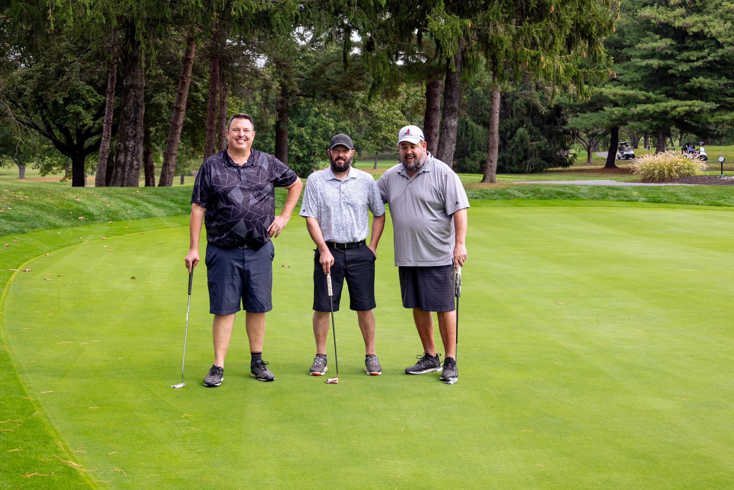 Three men standing on a golf course putting green, holding golf clubs, with trees and golf carts in the background.