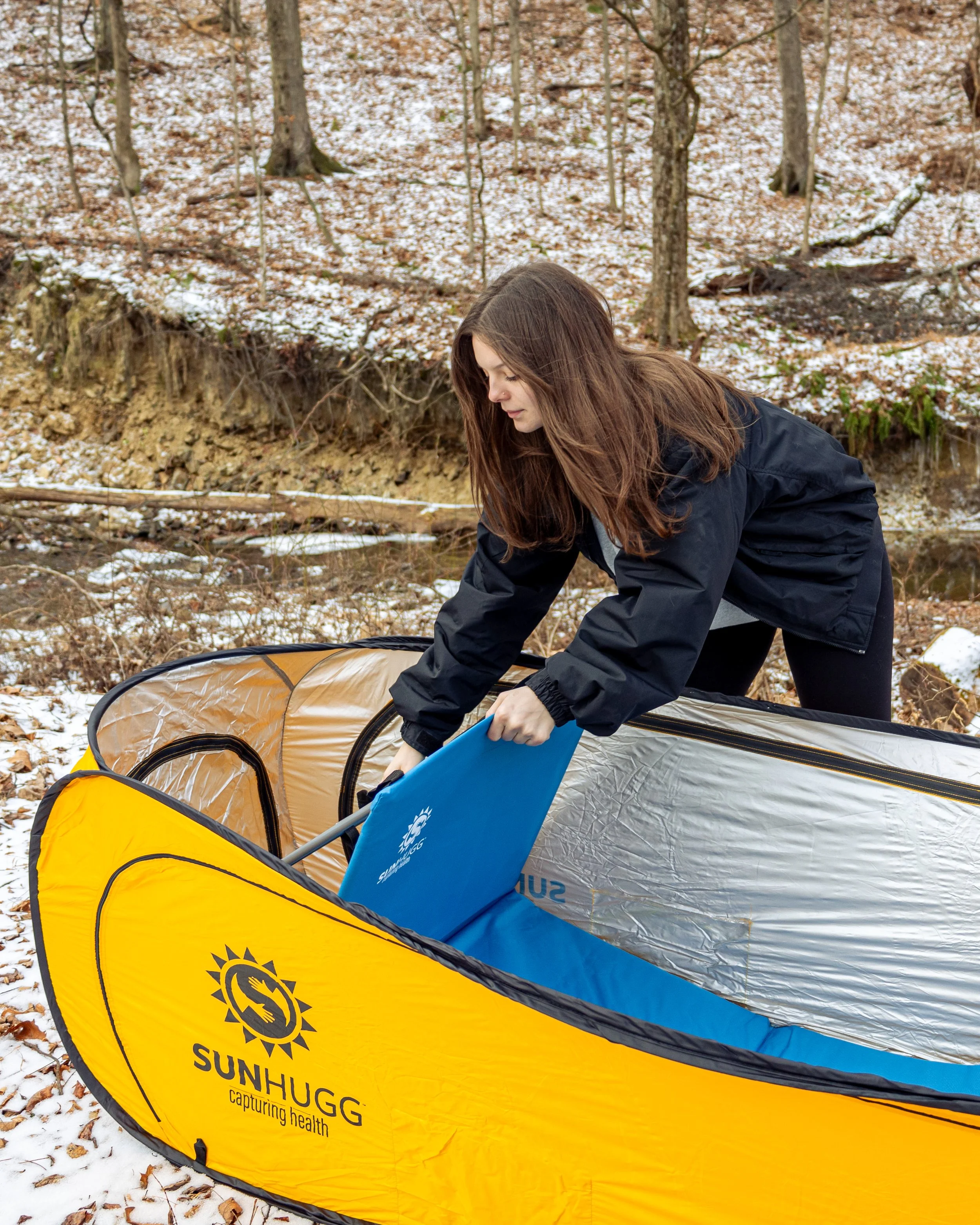A woman in an outdoor wooded area with snow on the ground, bending over to place a blue surfboard into a yellow Sunhugg portable shelter.