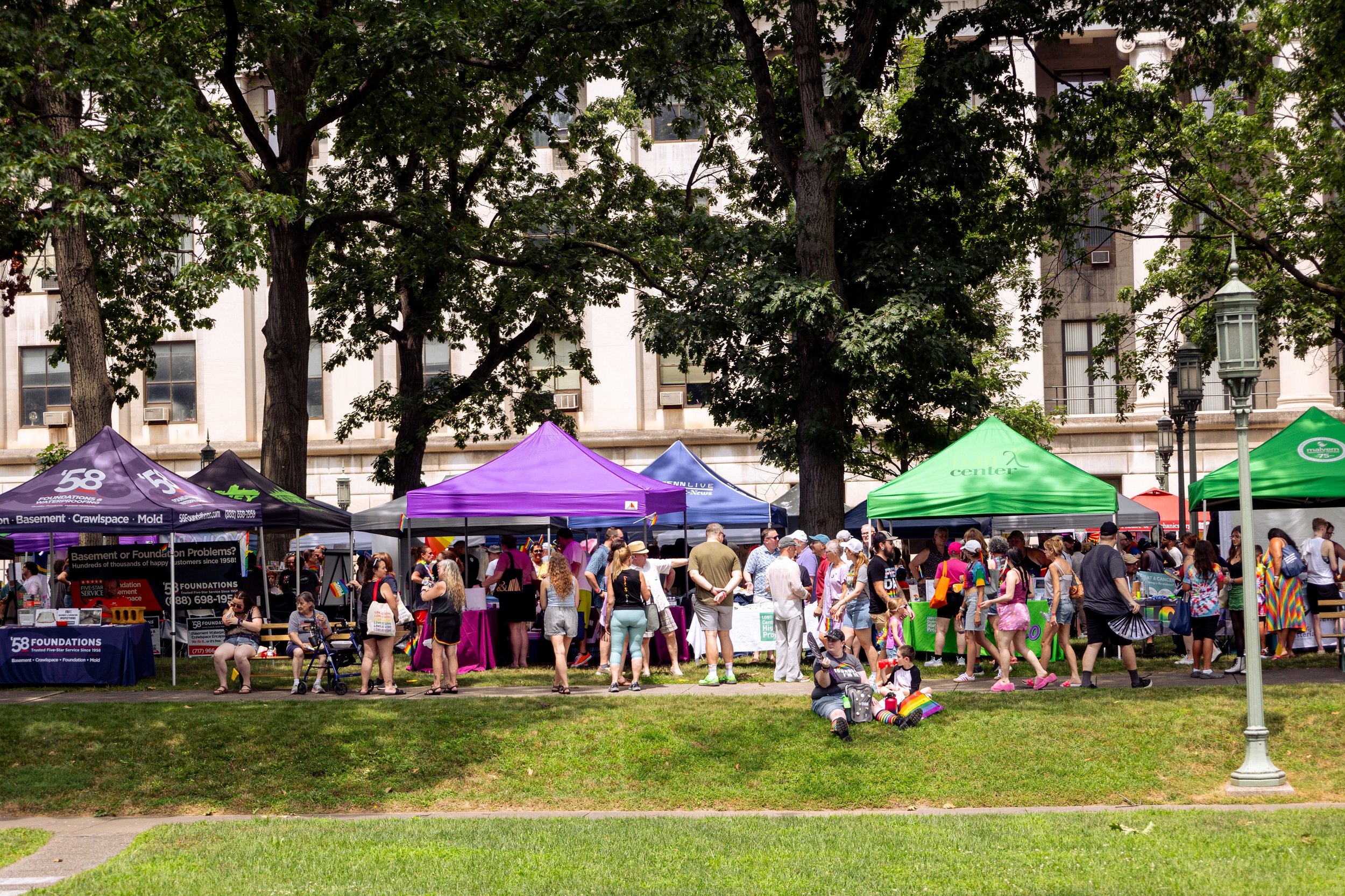 Crowd of people at an outdoor fair or market with colorful tents in a park setting, trees, and a large historic building with windows and air conditioning units in the background.