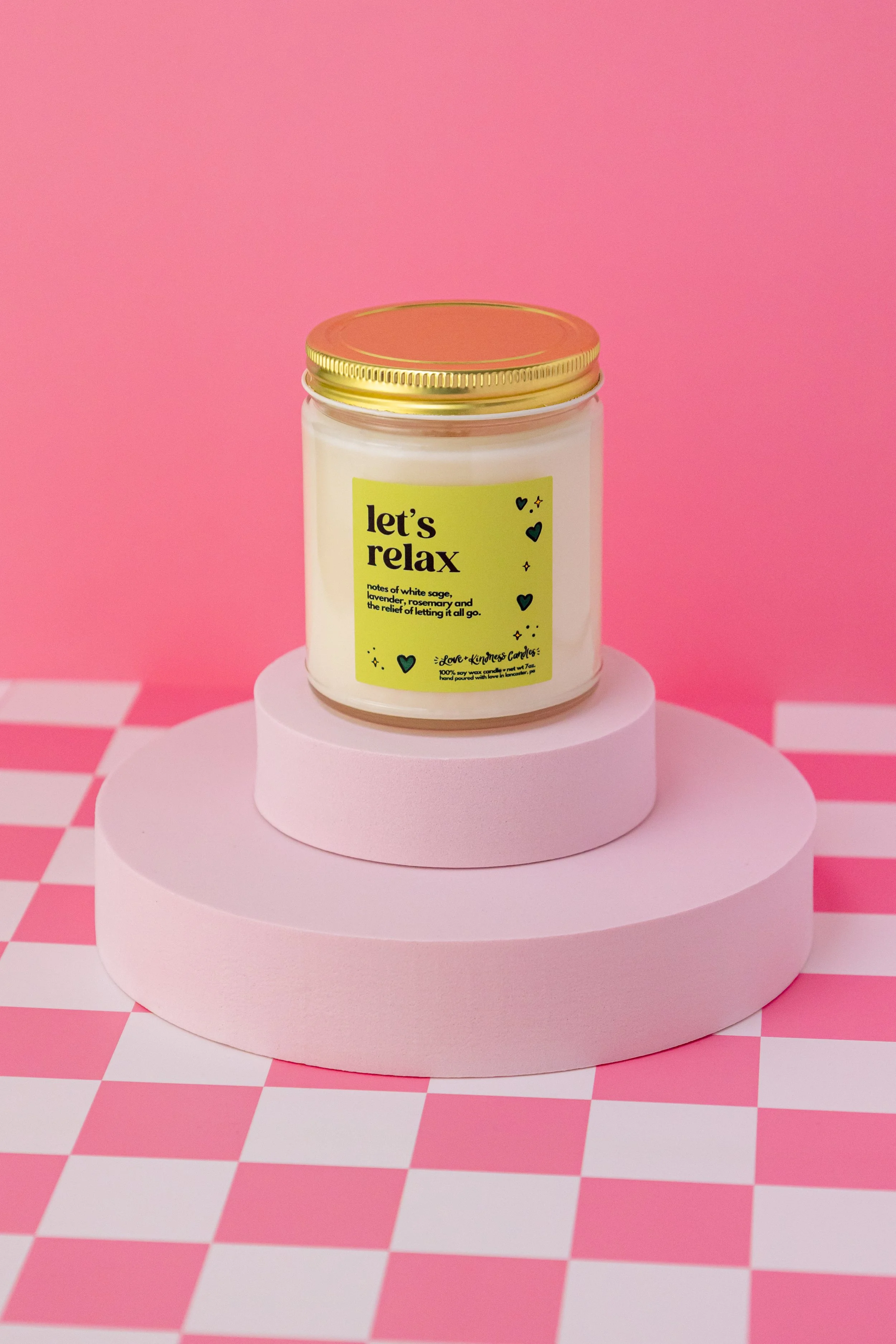 Love + Kindness Candles "Let's Relax" candle on cylindrical pink risers with a pink background and checkered white and pink. This product photography project focused on Love + Kindness Candles’ rebrand, creating clean, consistent visuals for product 