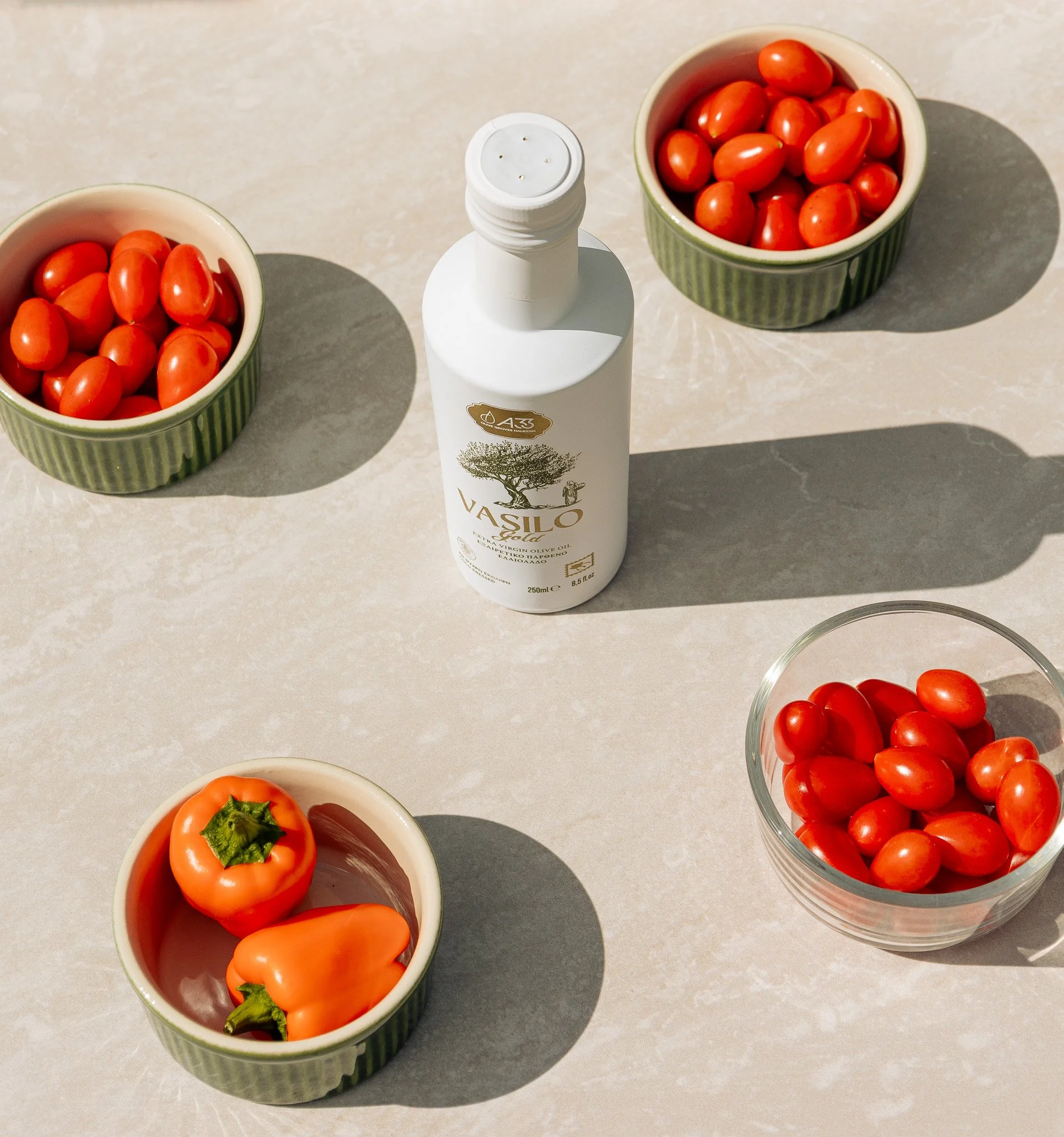 Bowl of cherry tomatoes, bowl of grape tomatoes, bowl of red and orange bell peppers, bottle of extra virgin olive oil on a light-colored surface, with shadows cast by the sunlight.