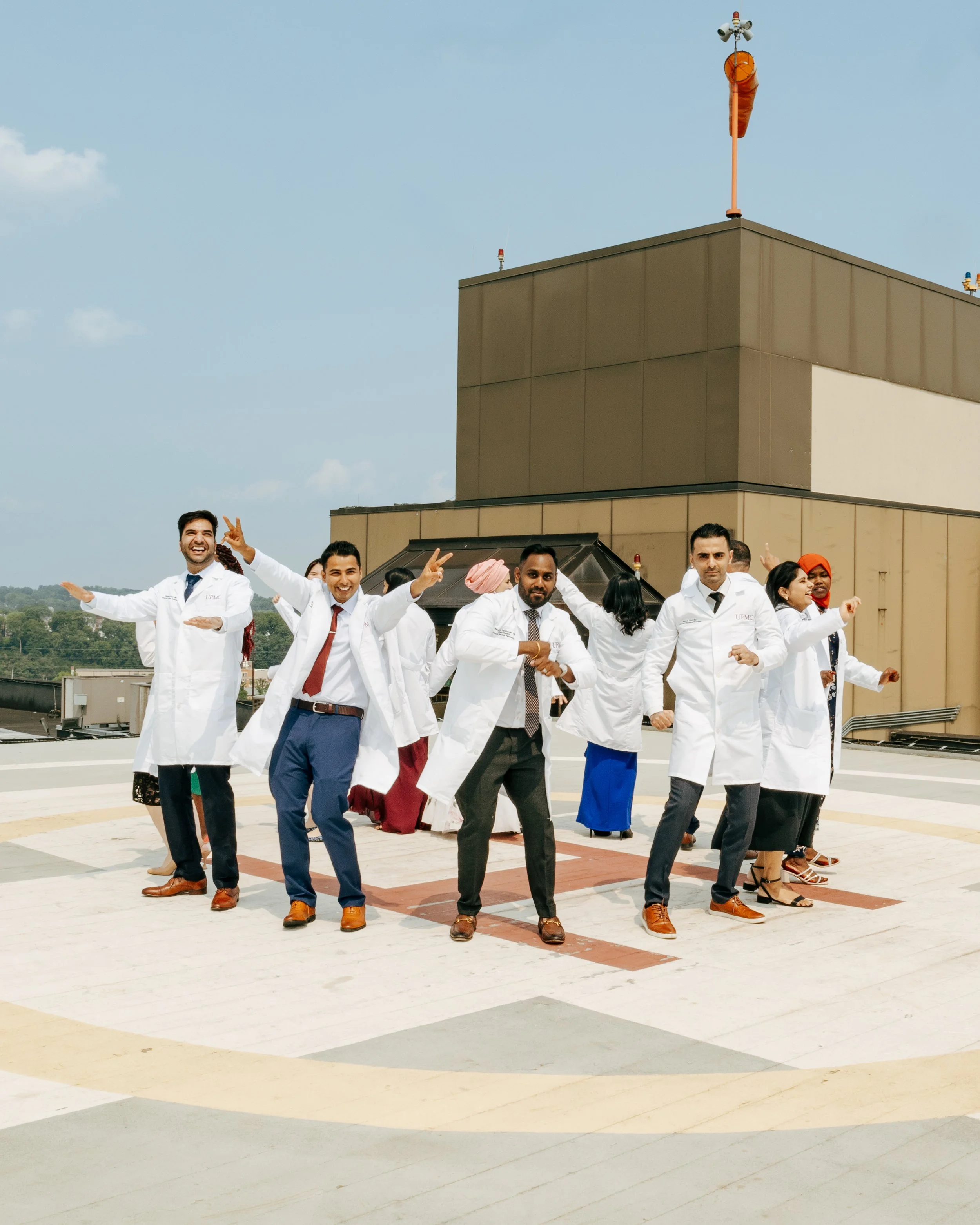 Group of healthcare professionals, mostly doctors in white coats, on a rooftop helipad, smiling and dancing in a celebratory manner.