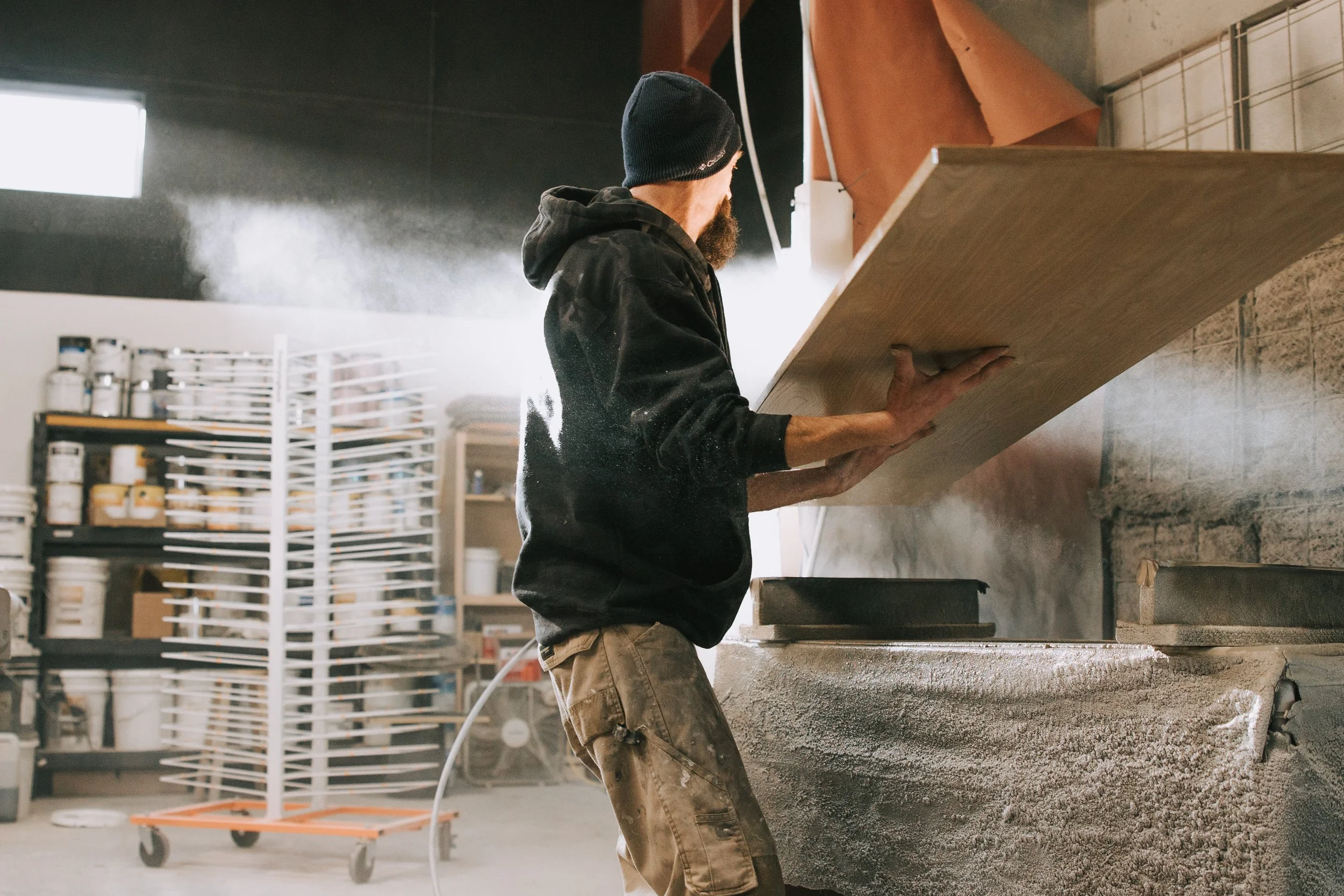 A man wearing a black hoodie, camouflage pants, and a navy beanie is sanding a large wooden board in a workshop with shelves of supplies in the background.