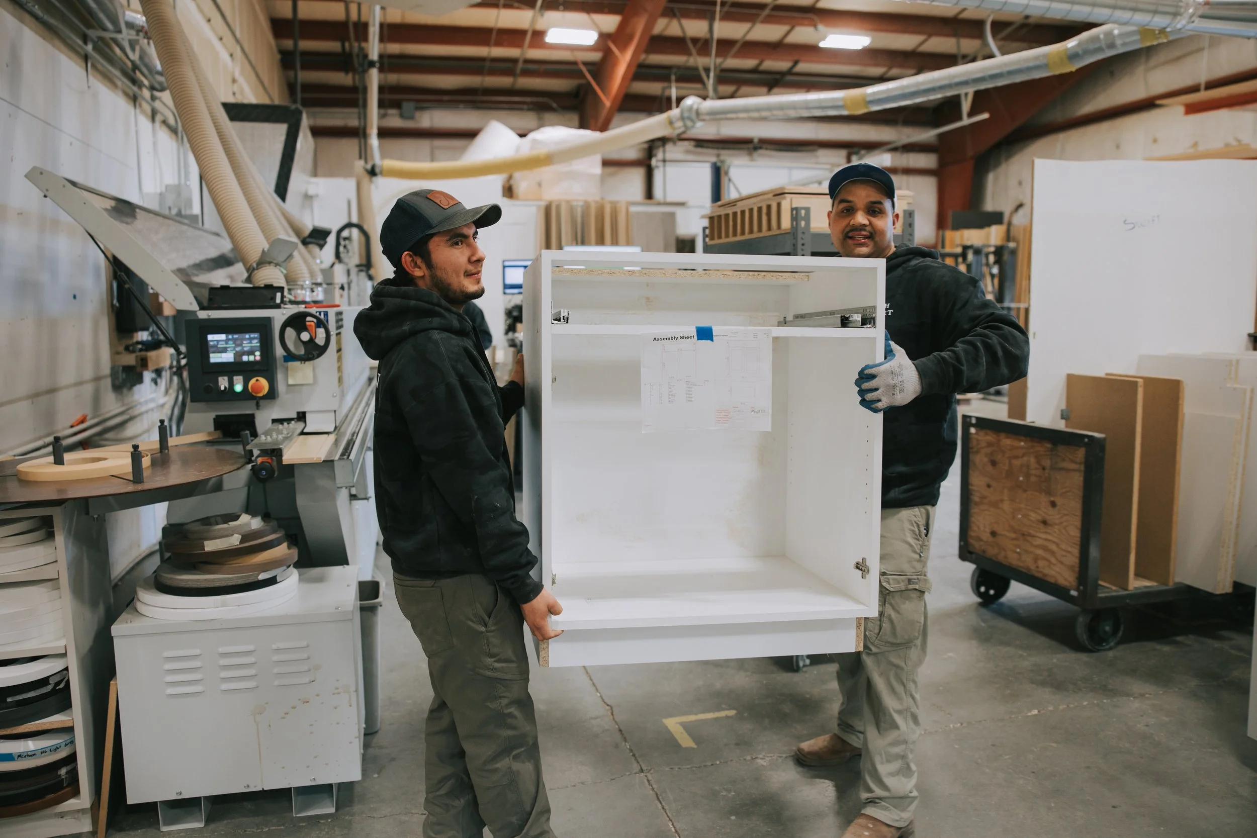Two workers inside a woodworking shop holding a white cabinet together, with woodworking tools and panels around.