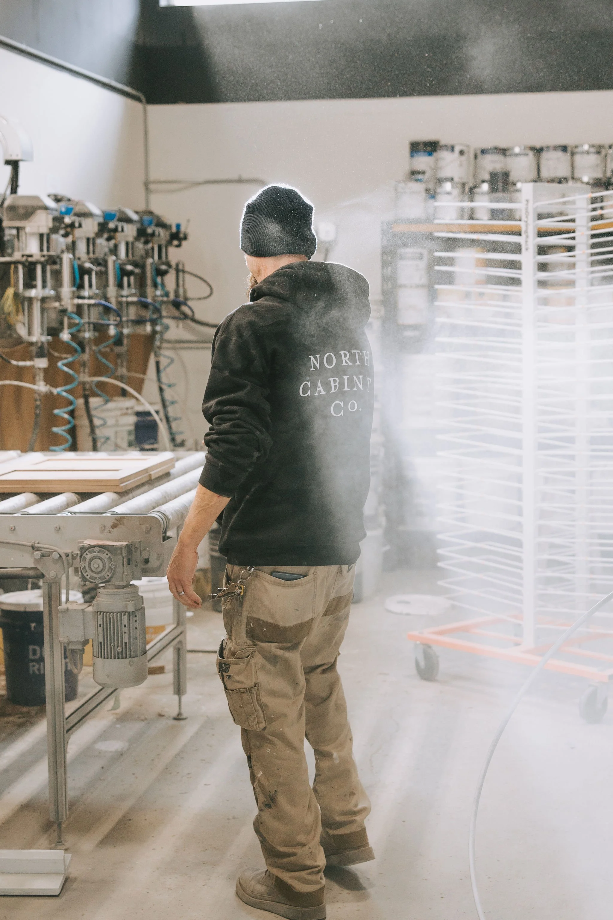 A man wearing a black hoodie and beige work pants inside a workshop, standing near a machine with smoke or vapor around him.