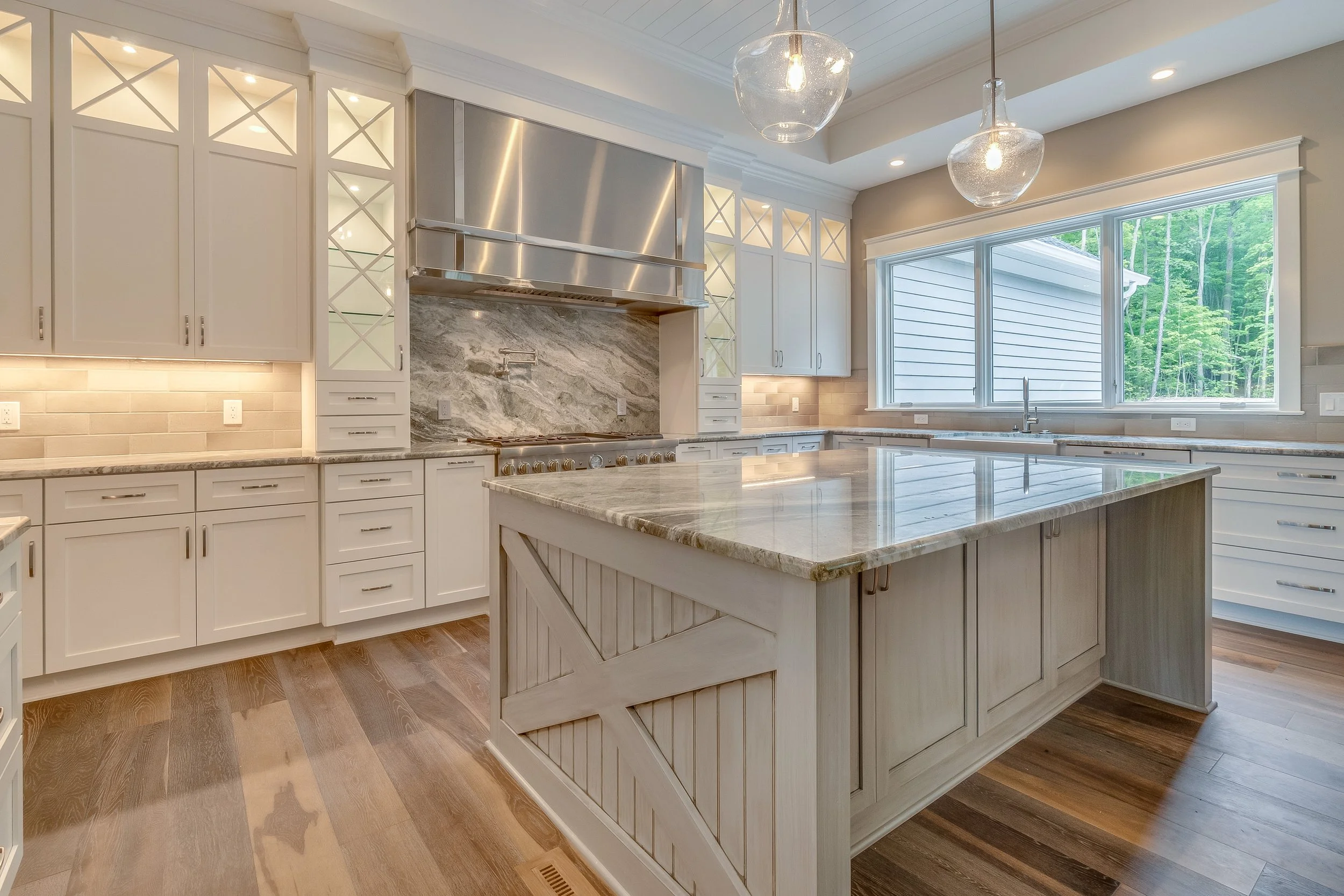 Modern kitchen with white cabinets, a large island with a marble countertop, stainless steel range hood, and wood flooring. Large window overlooking green trees.