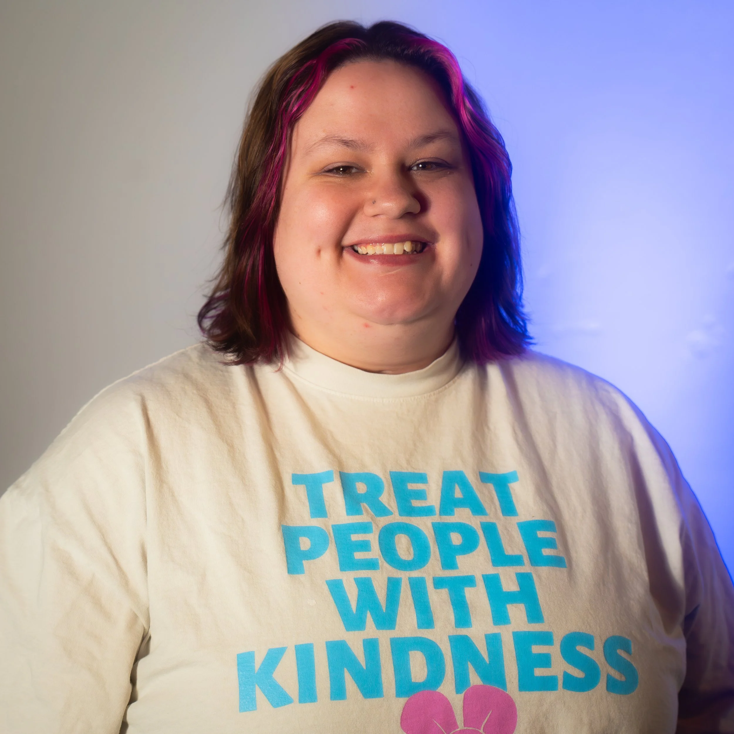 A smiling woman with shoulder-length dark hair with pink highlights, wearing a beige t-shirt with blue text reading 'Treat People With Kindness' and a pink heart illustration.
