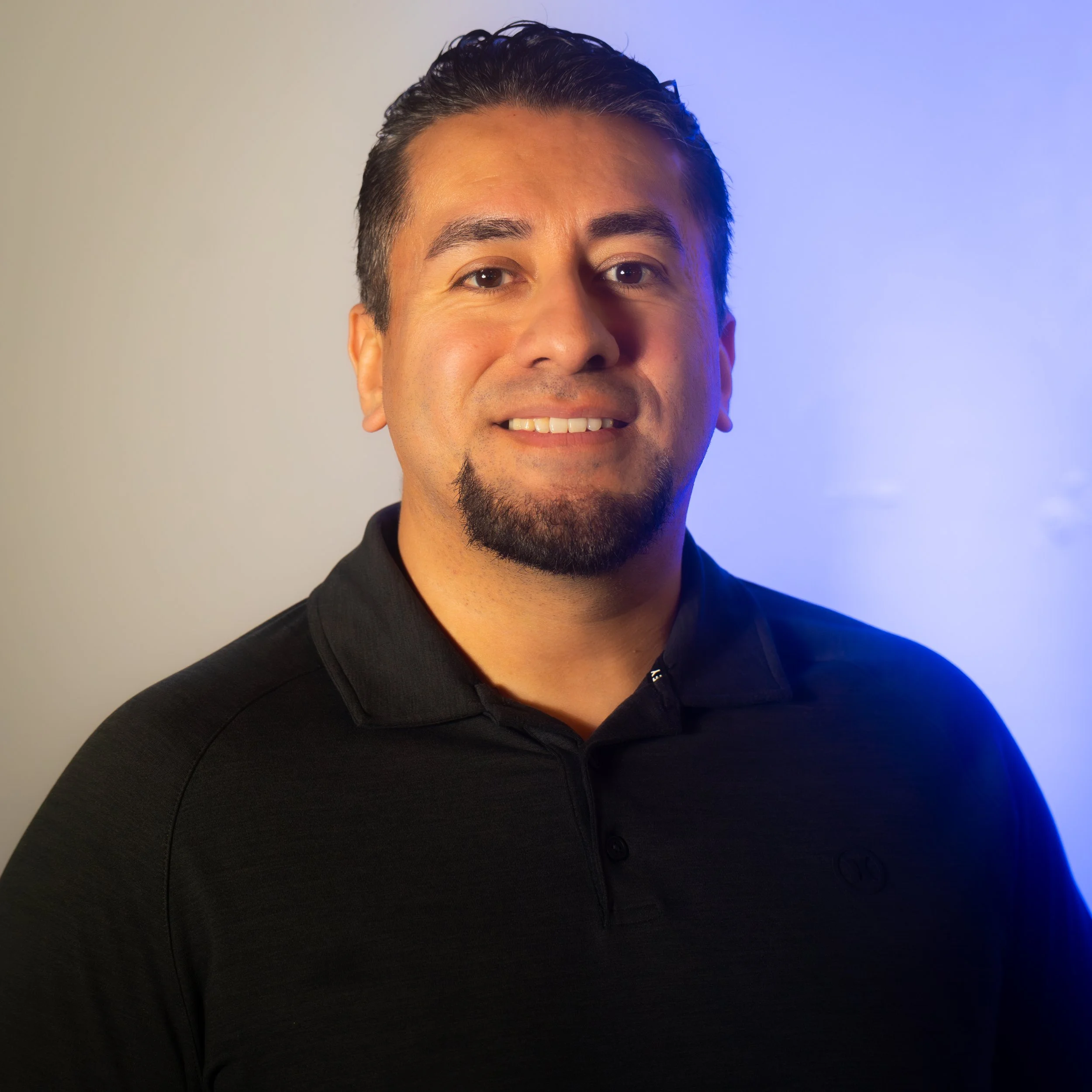 A man with short dark hair, a goatee, and a goatee mustache smiling, wearing a black collared shirt, standing indoors with a neutral background and blue lighting on the right side.