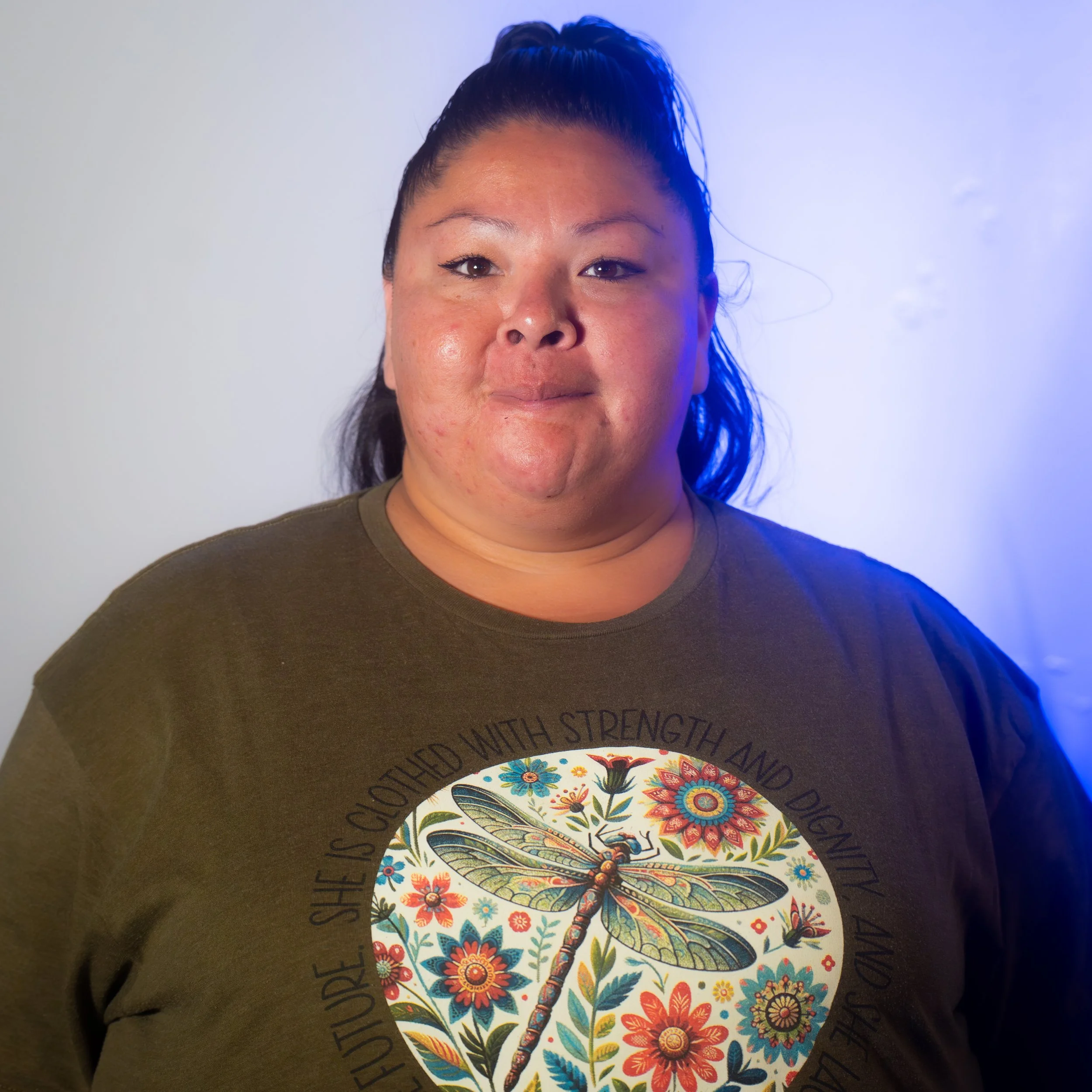 A person with long dark hair tied back, wearing a dark green T-shirt with a dragonfly and floral design on the front, standing against a plain light background.