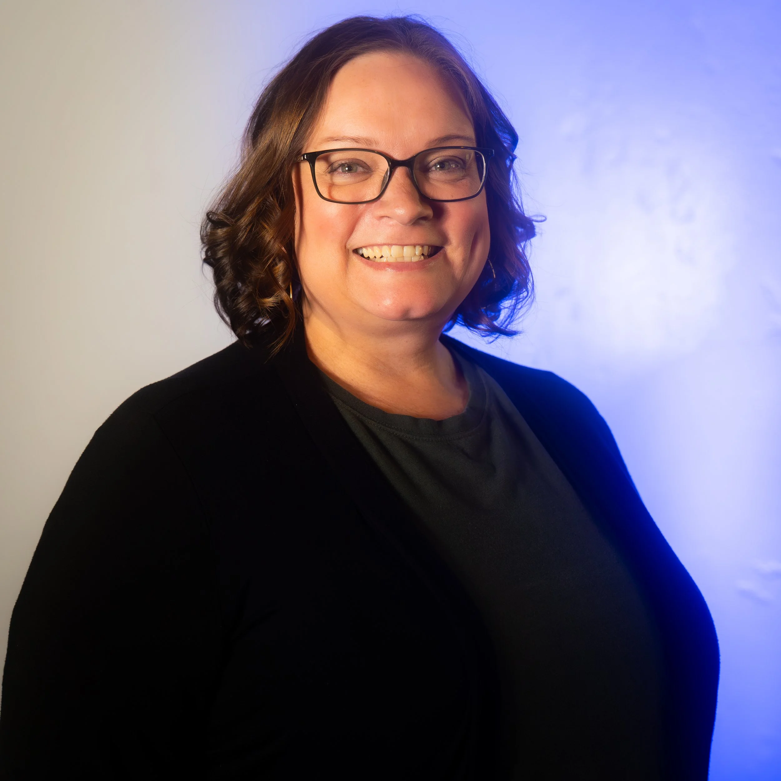A smiling woman with glasses, shoulder-length wavy brown hair, wearing a black jacket and a dark shirt, standing against a background with a gradient from white to blue.