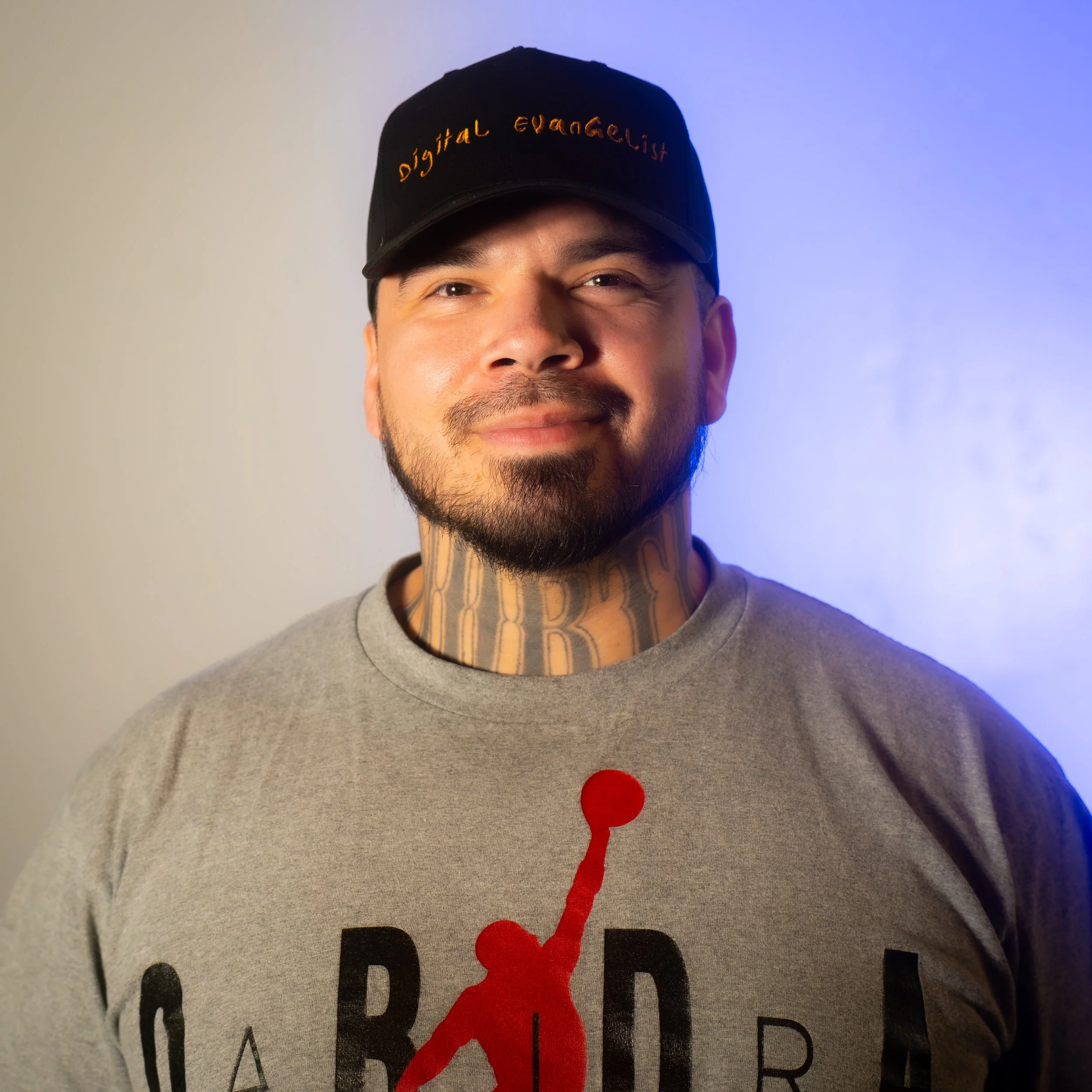 Man wearing a black cap with orange embroidered text, gray sweatshirt with a red and black graphic, and tattoos on his neck, smiling in front of a neutral and blue background.