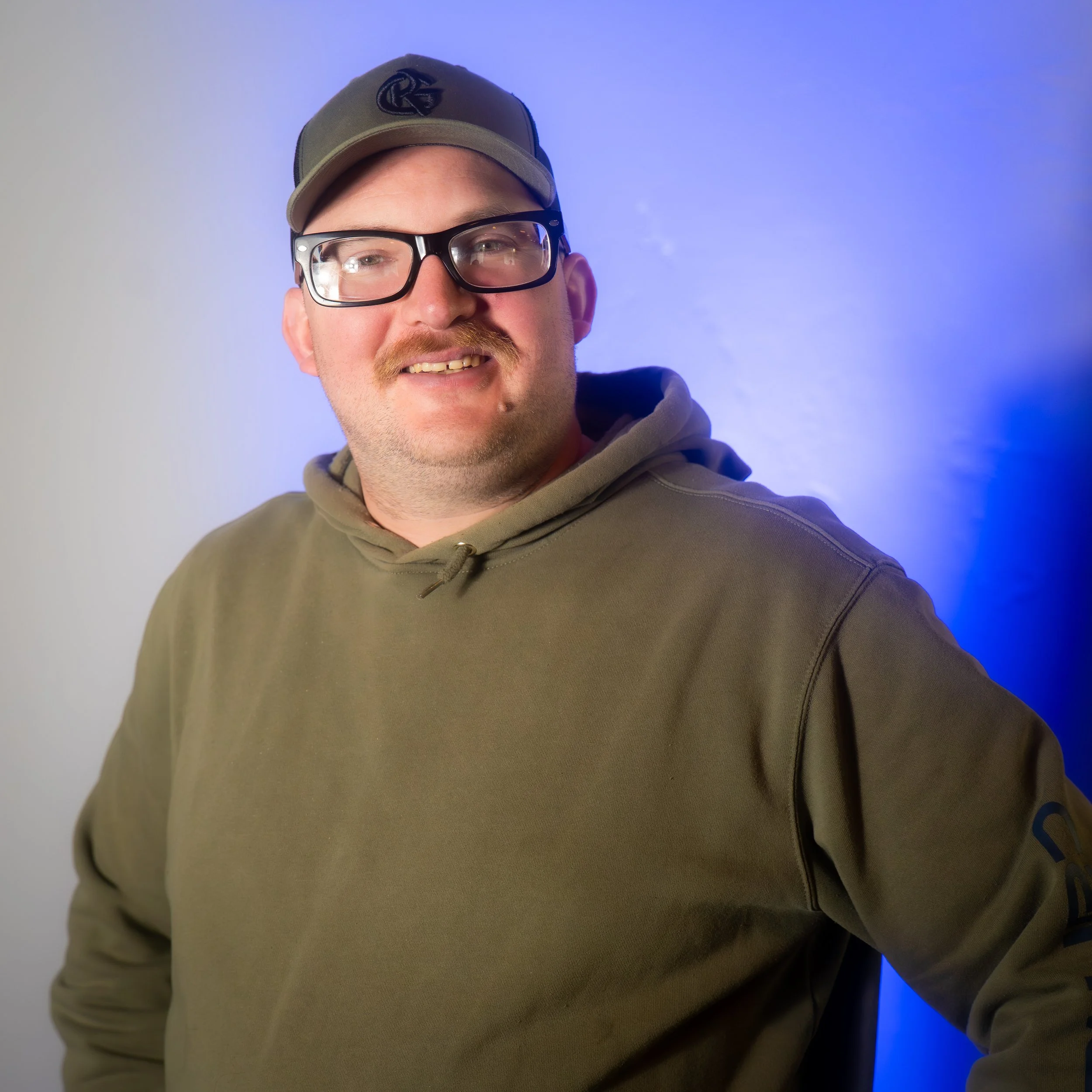A man wearing glasses, a baseball cap, and a green hoodie, smiling and standing against a blue and gray background.