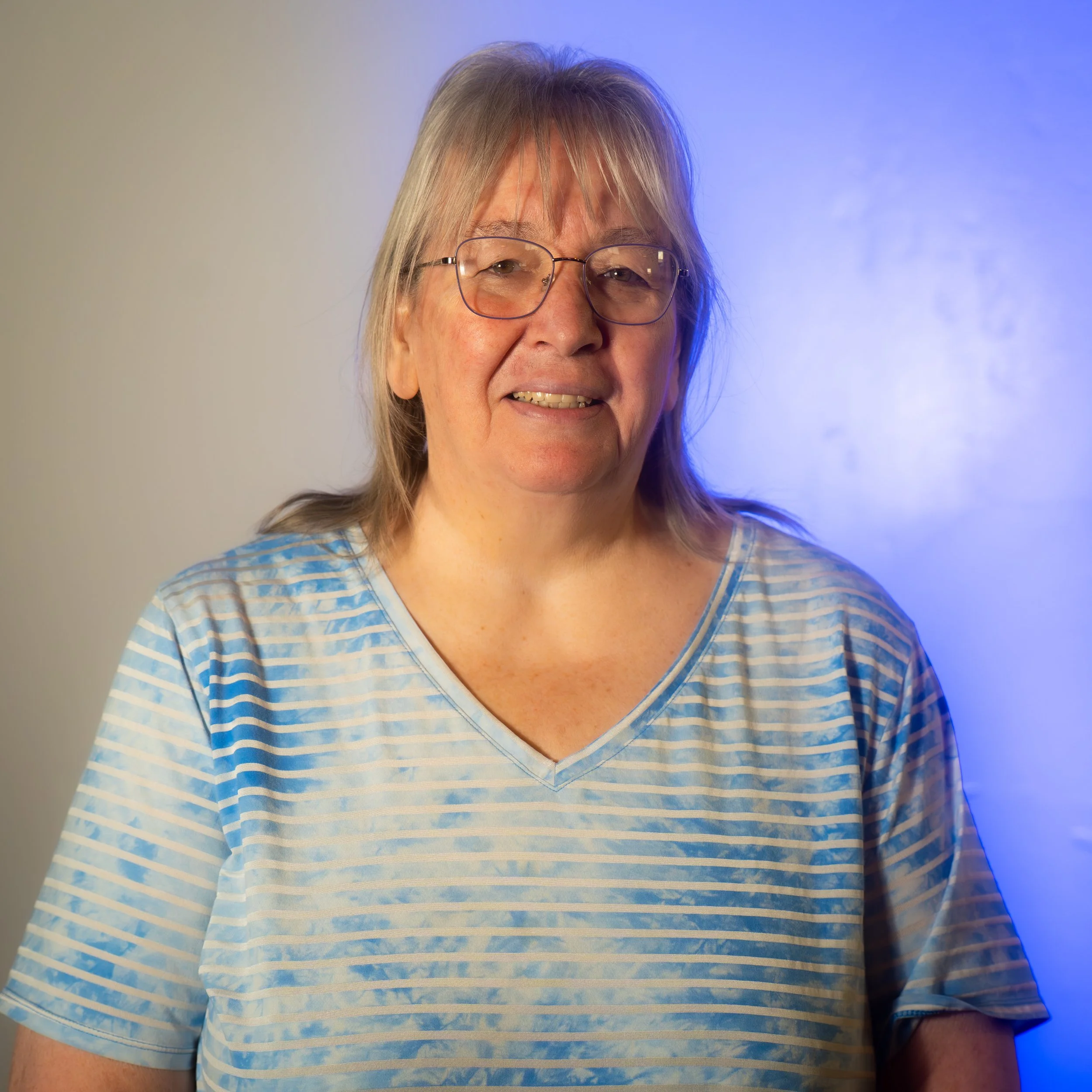 A smiling older woman wearing glasses and a blue and white striped V-neck T-shirt standing against a neutral background with a subtle blue lighting effect.