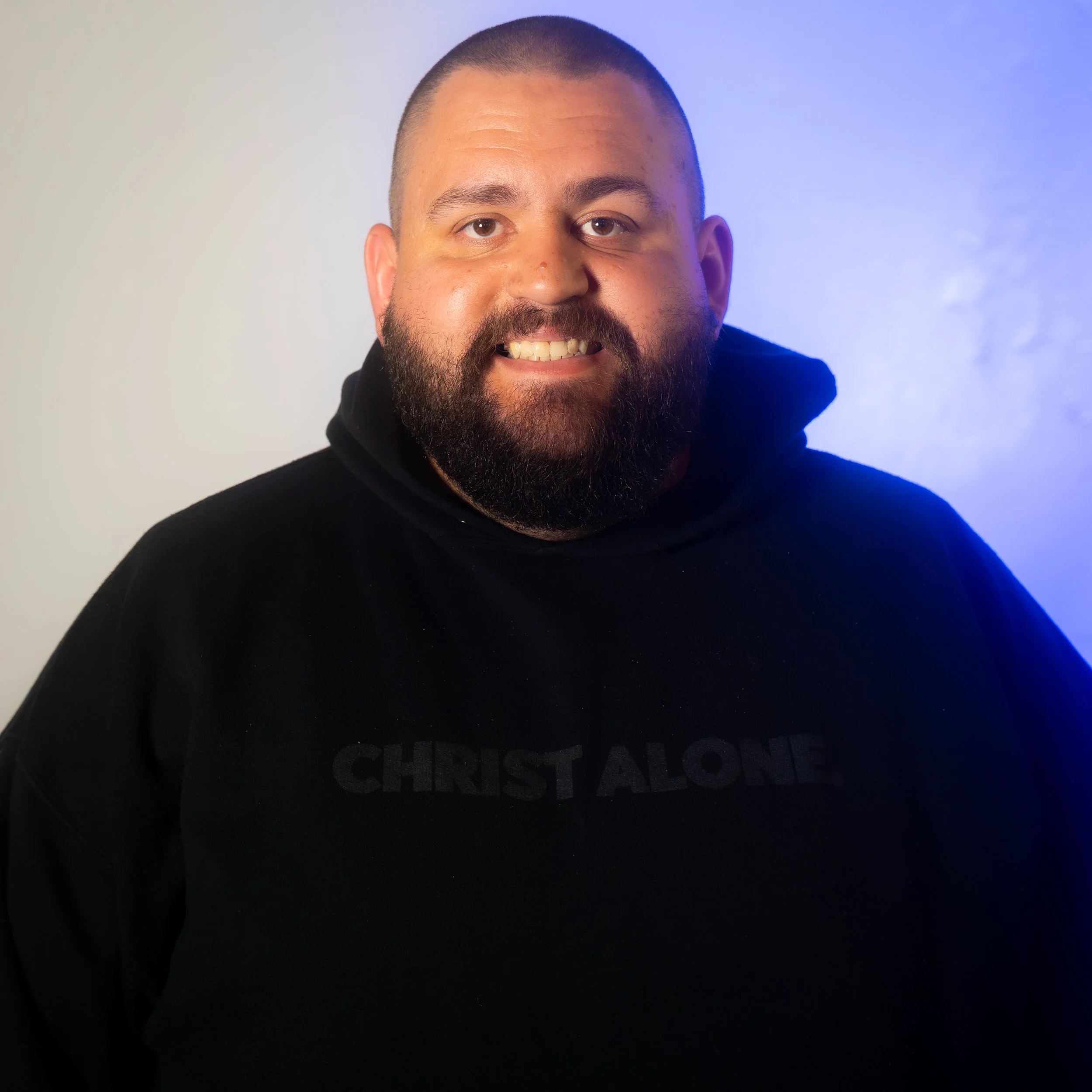 A man with a beard and short hair smiling in front of a gradient background wearing a black hoodie with 'CHRIST ALONE' printed on it.