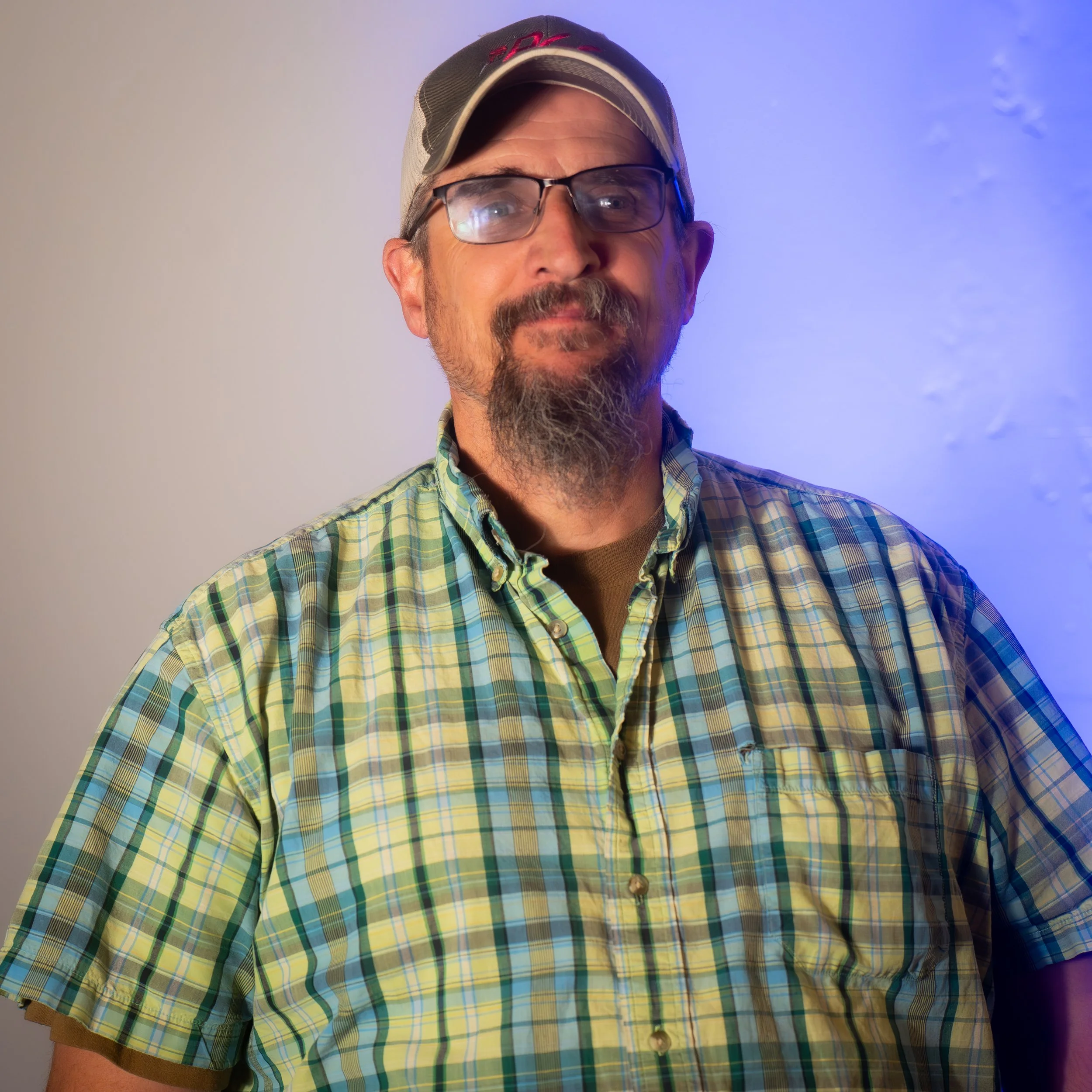 A man with glasses and a beard wearing a baseball cap and a plaid short-sleeve shirt, standing in front of a background with gradient lighting.