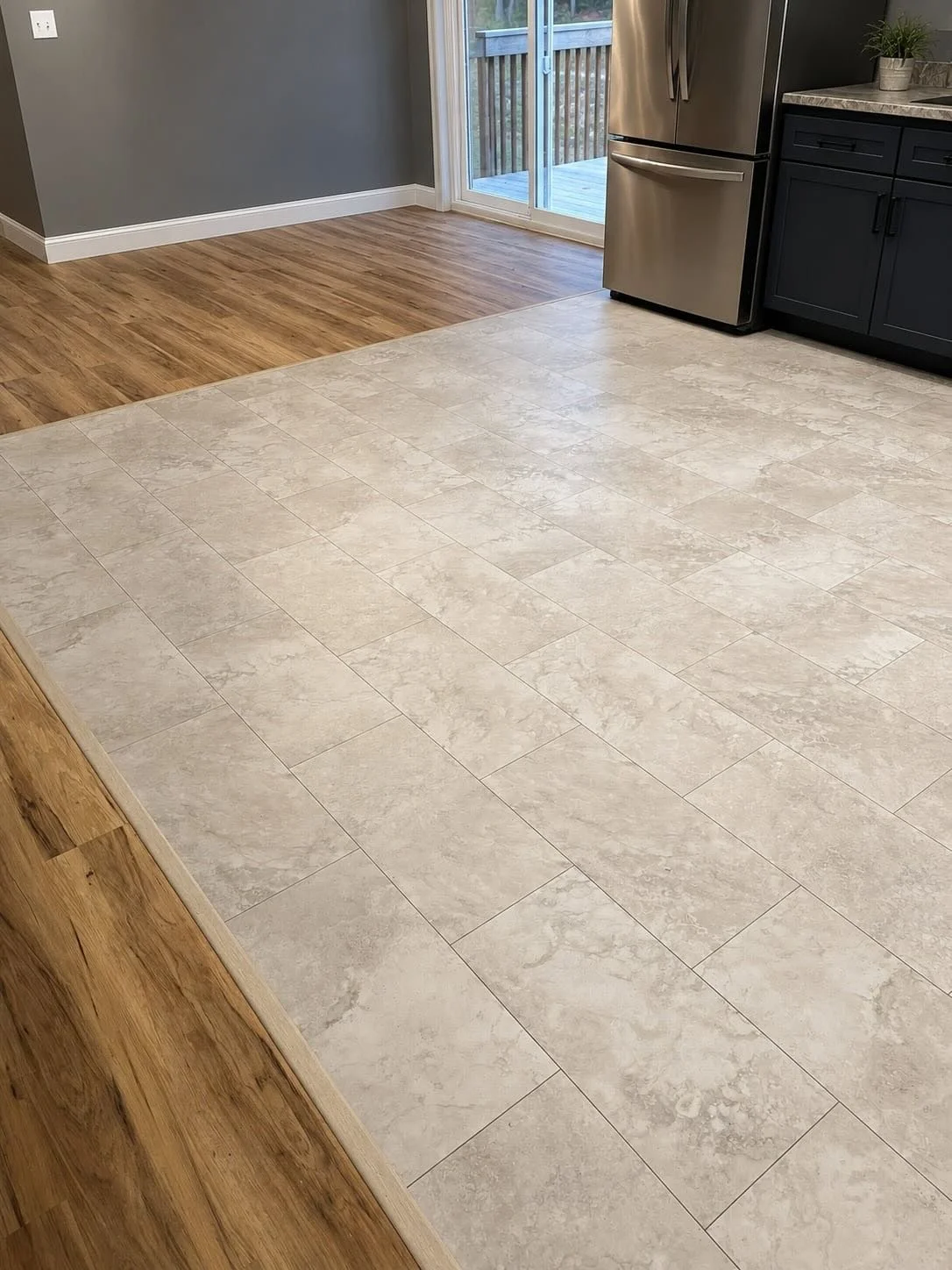 Modern kitchen featuring large-format beige stone-look luxury vinyl tile flooring installed in a staggered pattern, dark shaker cabinets, stainless steel appliances, and adjacent wood-look flooring in an open-concept layout.
