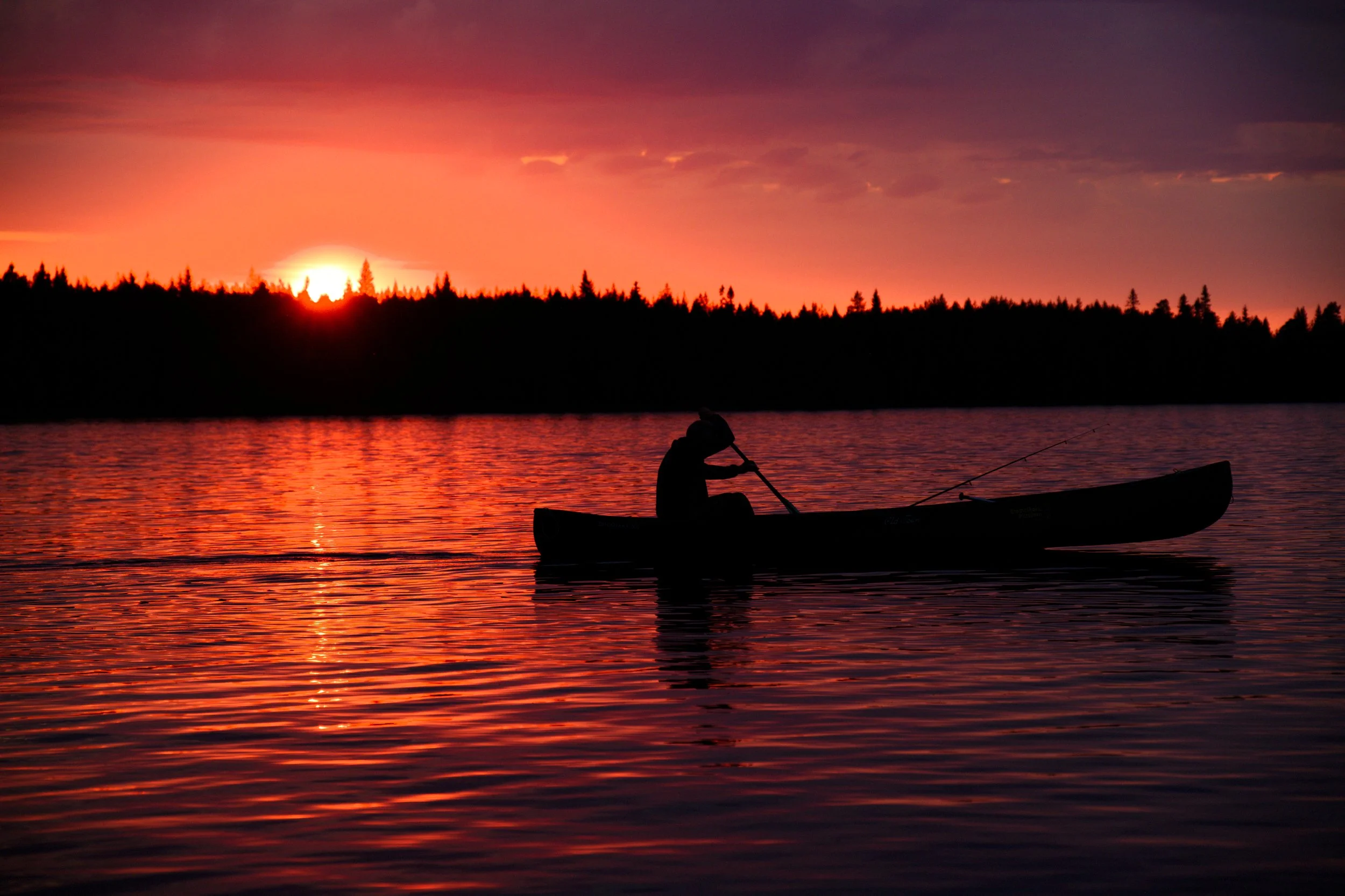 A person canoeing and fishing on a lake in Finland at sunset, with the silhouette of trees in the background, and the red, orange and purple colours of the setting sun