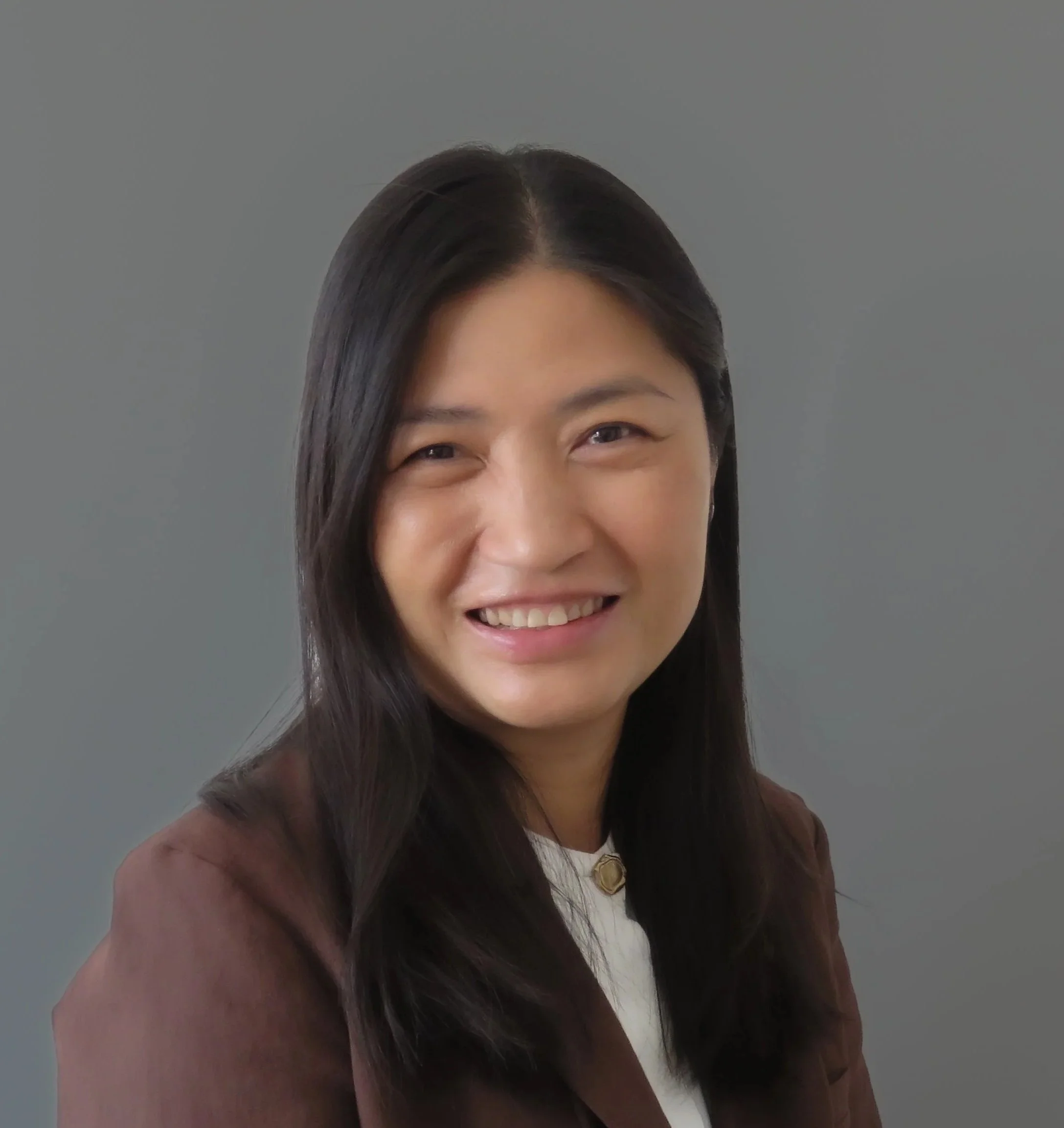 A woman smiling with straight black hair, wearing a brown jacket and a white top, against a plain gray background.