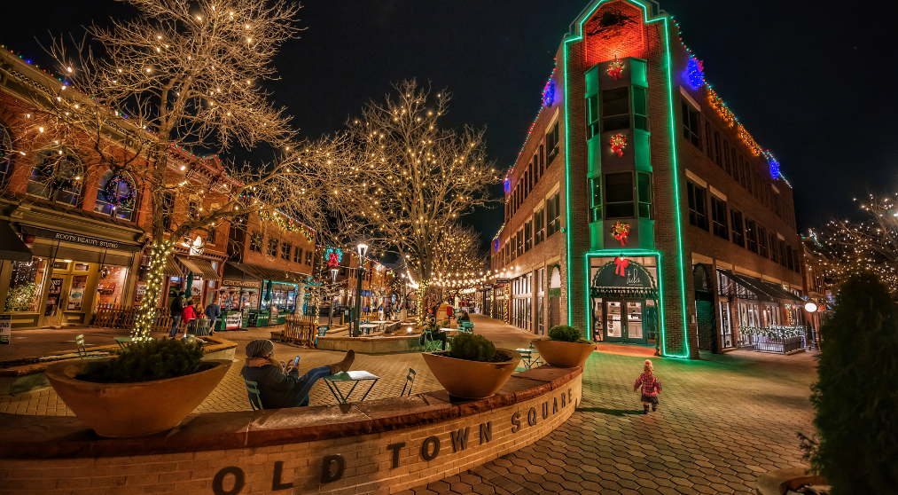 Festively decorated Old Town Square at night with lights on trees and buildings, people sitting and walking, child in foreground.