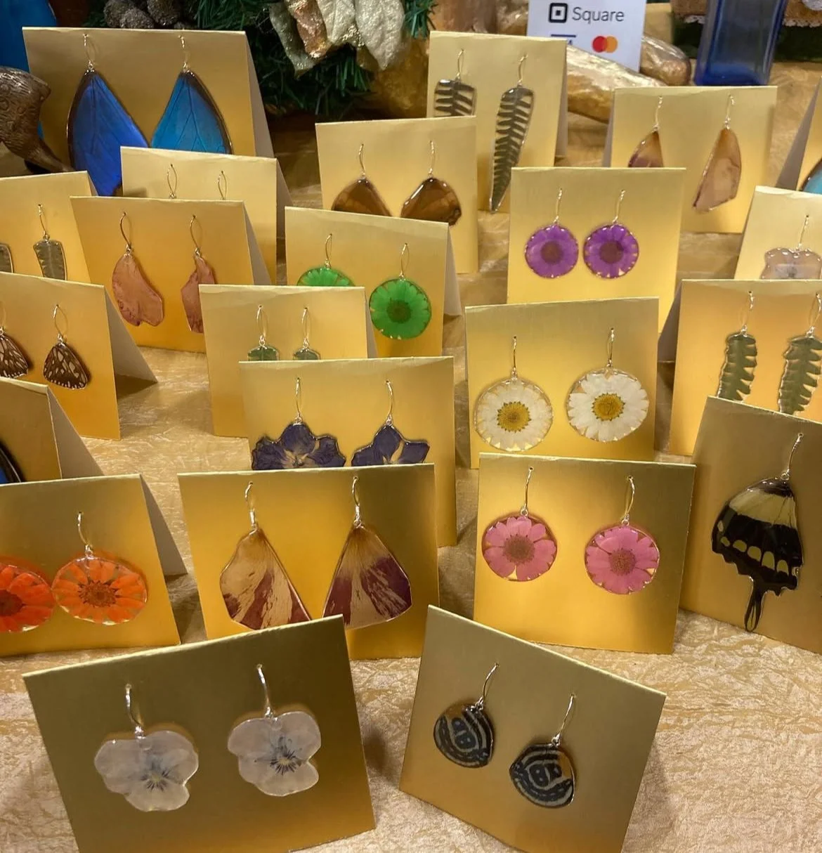 Display of earrings with natural designs on gold cards, featuring butterflies, flowers, and leaves, at a market stall.