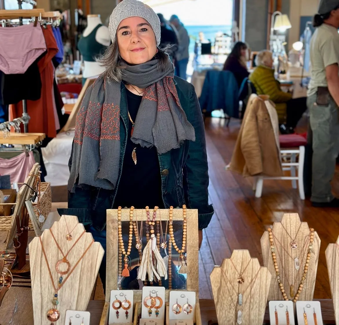 Woman in winter clothing stands behind a display of handmade jewelry at a market.