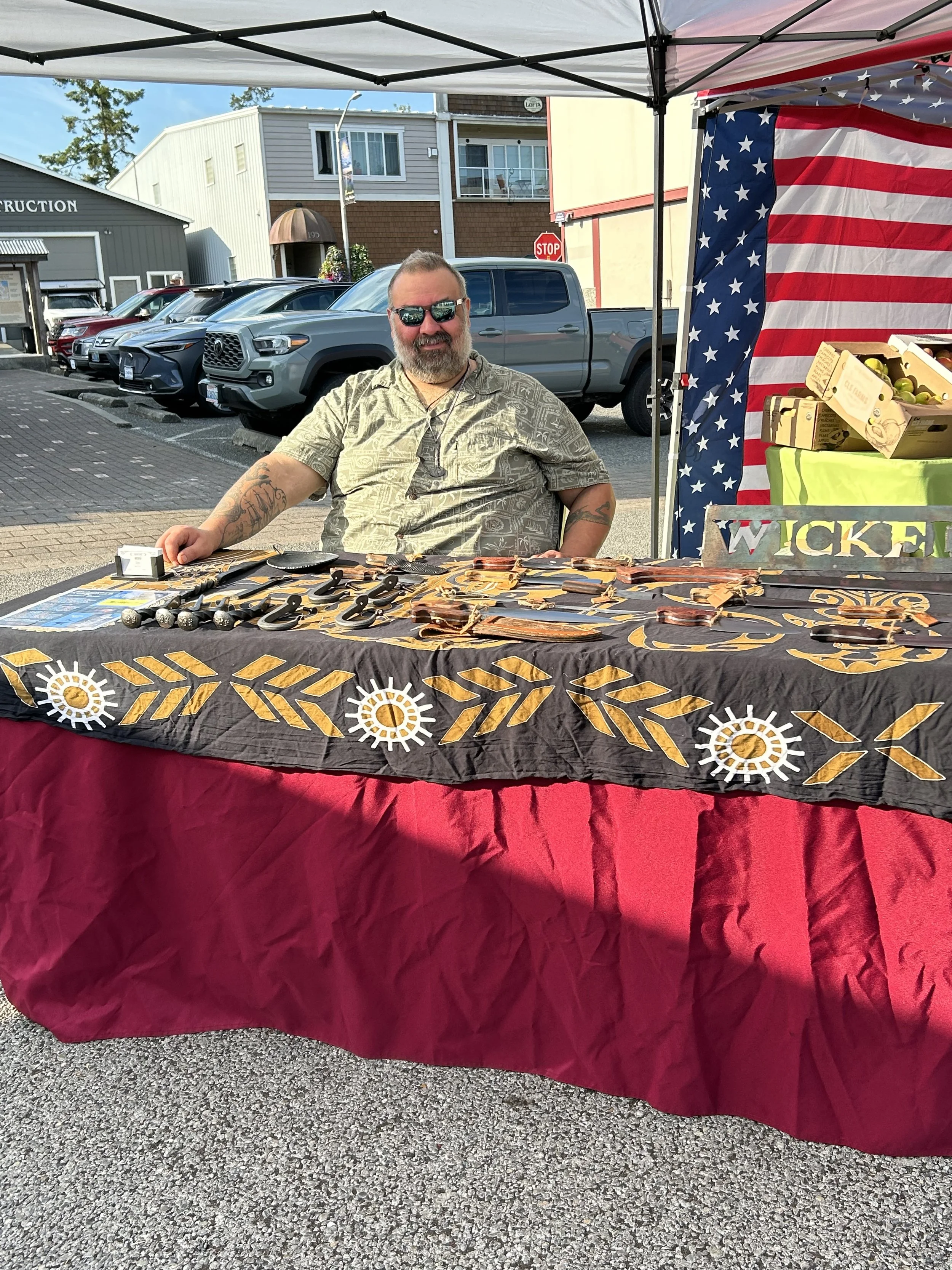 Man sitting at outdoor market stall with various handcrafted items on display, American flag, parked trucks, and building in background.