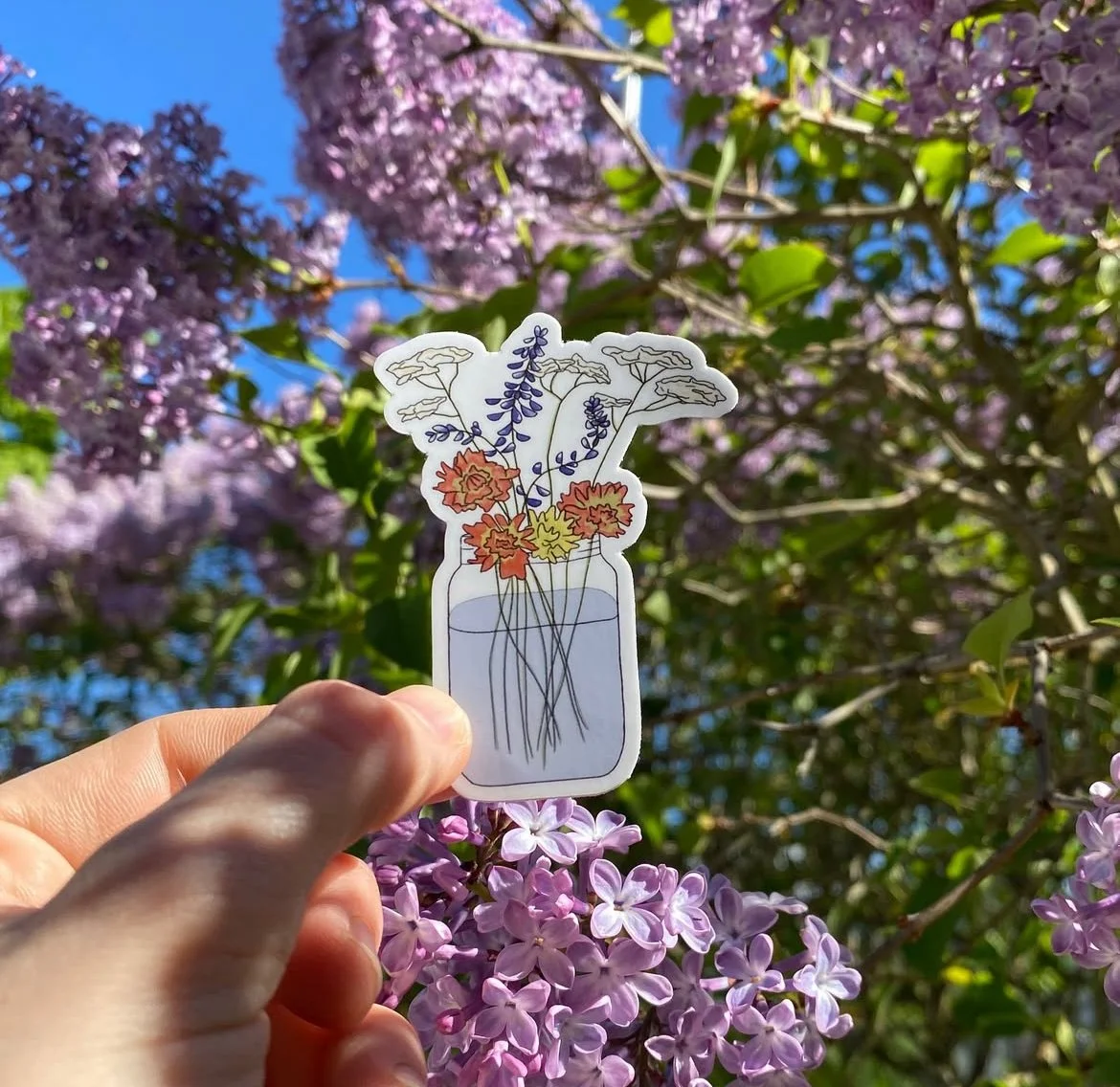 Hand holding a floral illustration in front of blooming lilac flowers and green leaves against a blue sky.