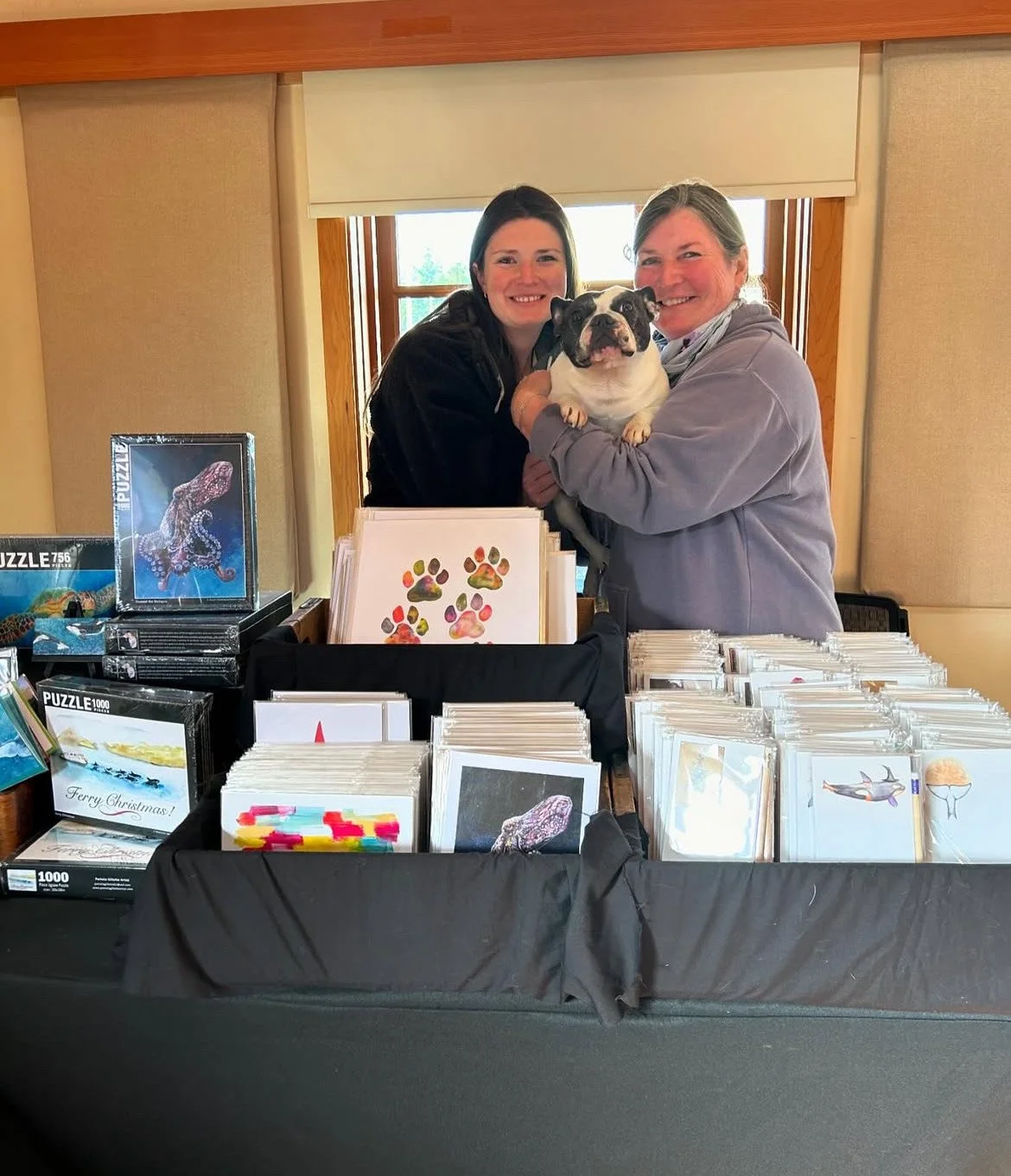 Two people smiling with a dog at a display table featuring puzzles and greeting cards.
