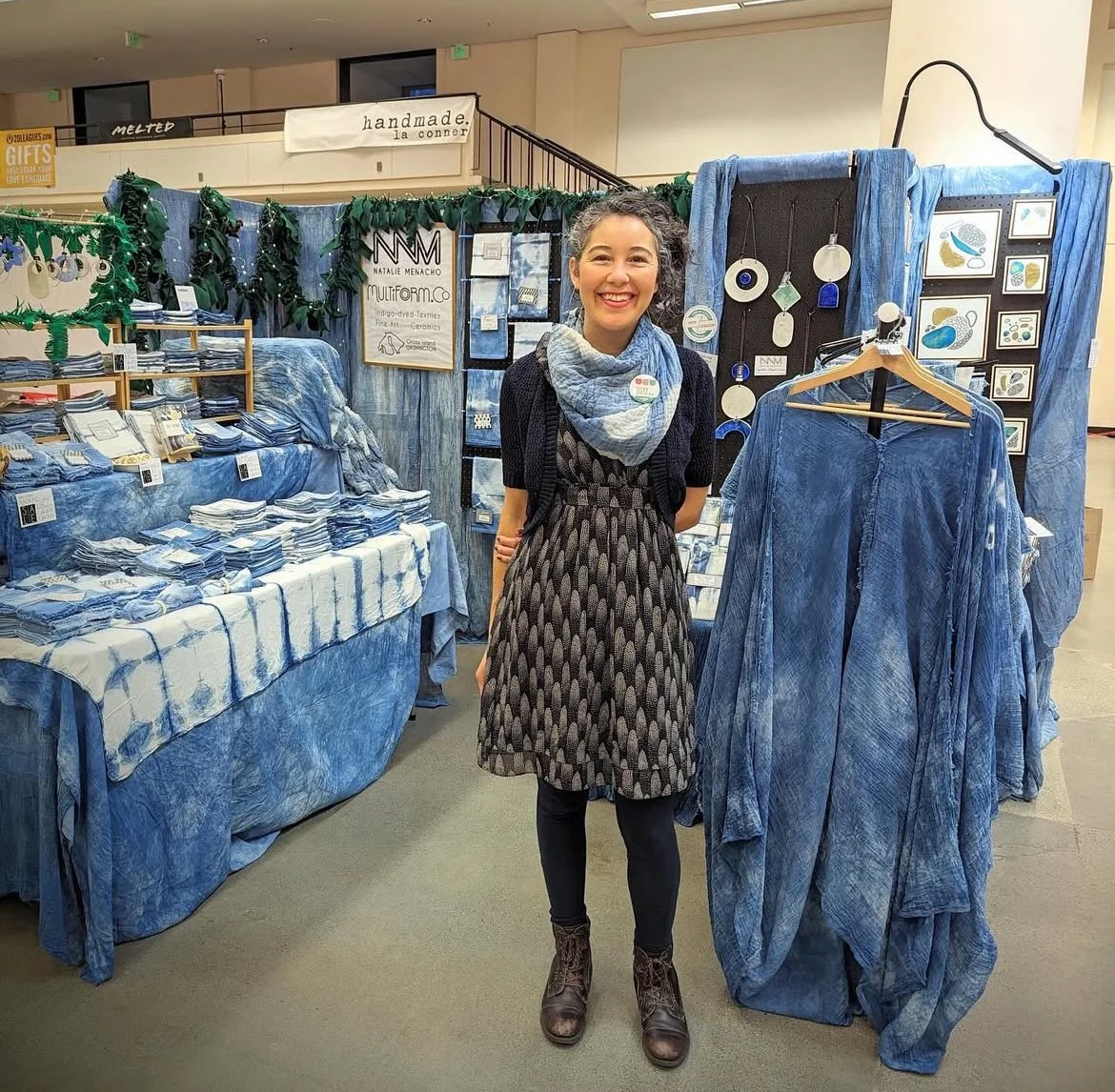 Woman standing in front of a textile booth showcasing blue-dyed fabrics and garments at a craft fair.