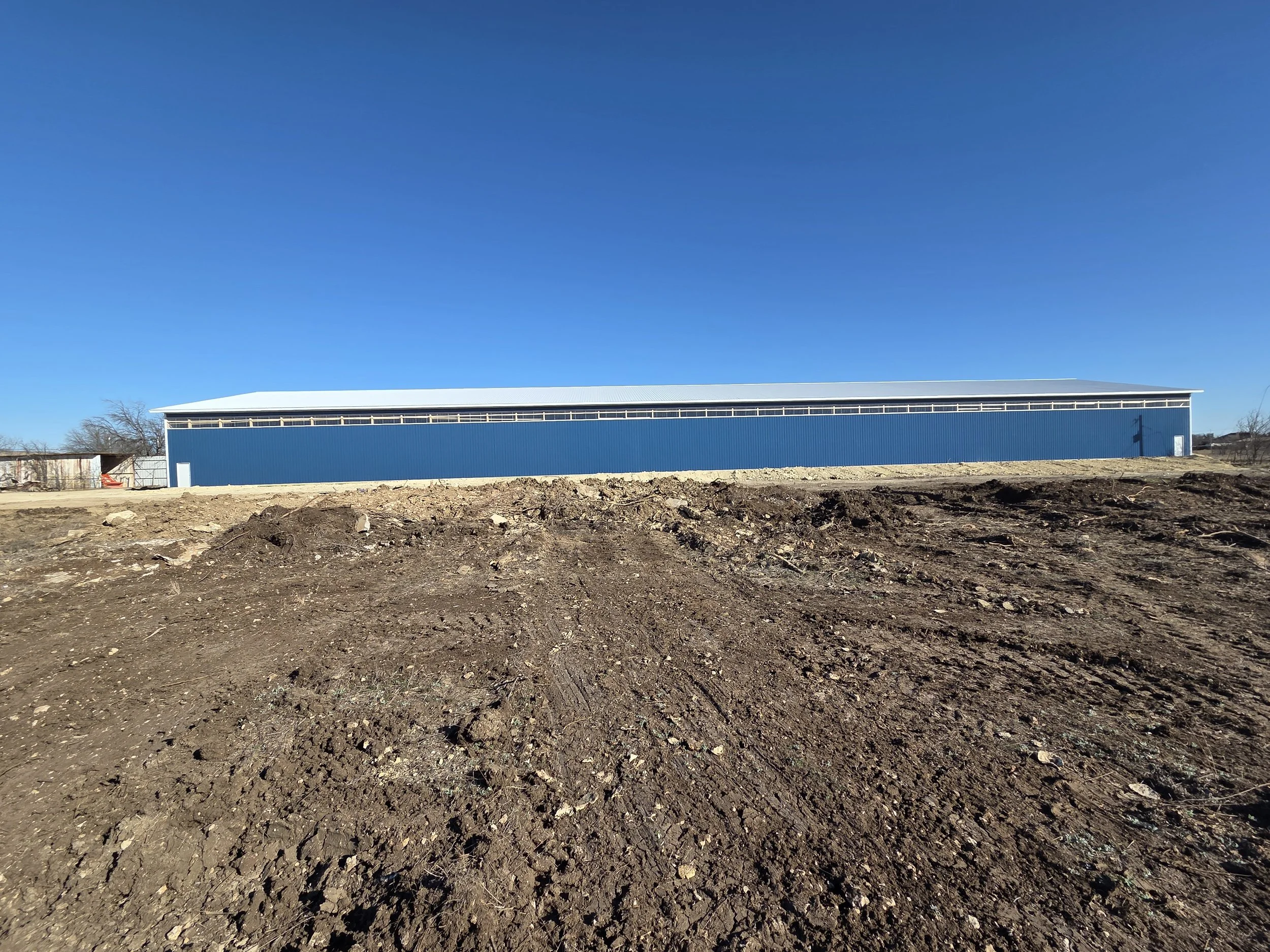 A long blue metal warehouse or storage building under a clear blue sky, with uneven dirt ground in the foreground.