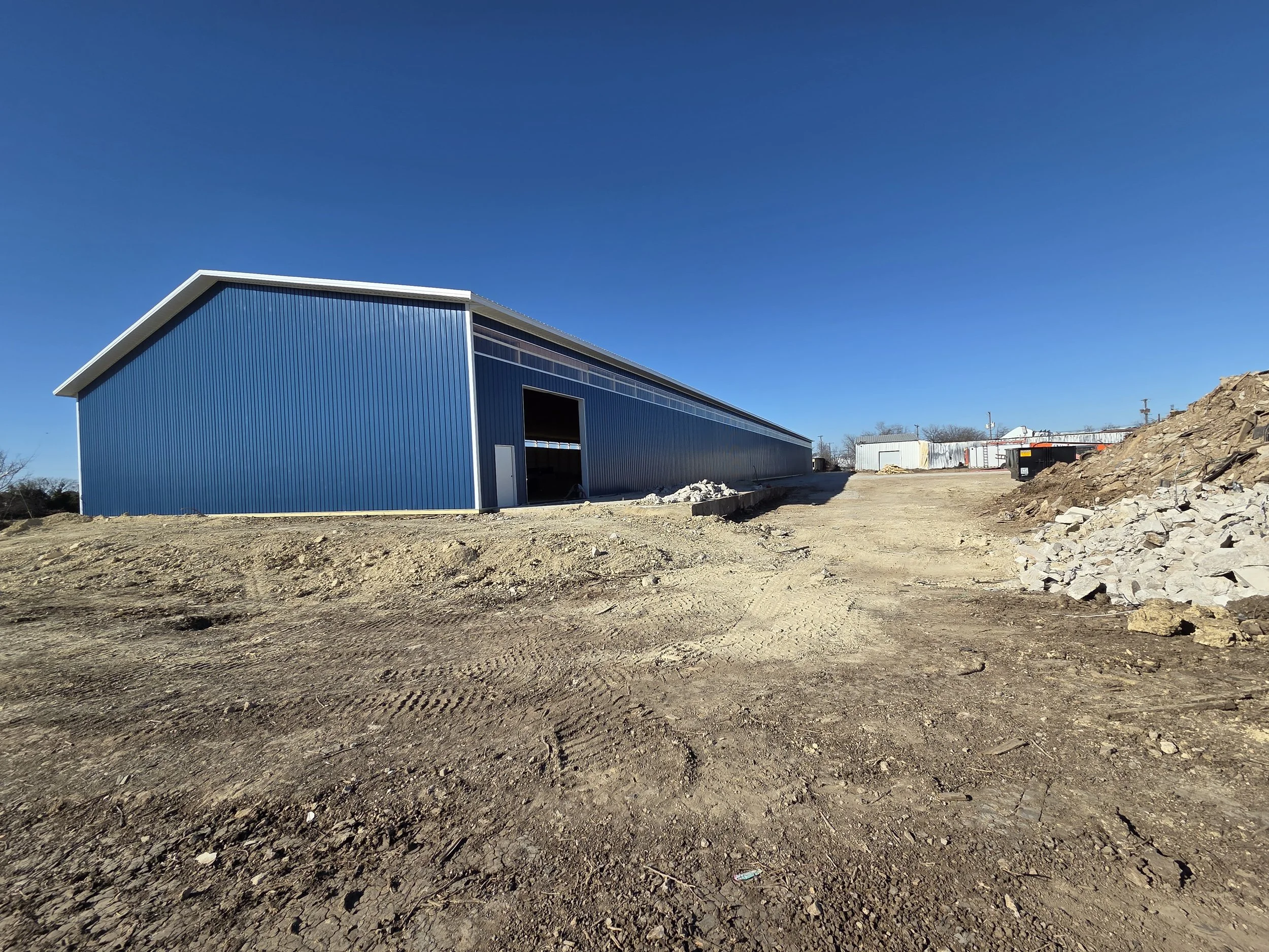 A large blue metal warehouse under construction on a dirt lot with clear blue sky in the background.