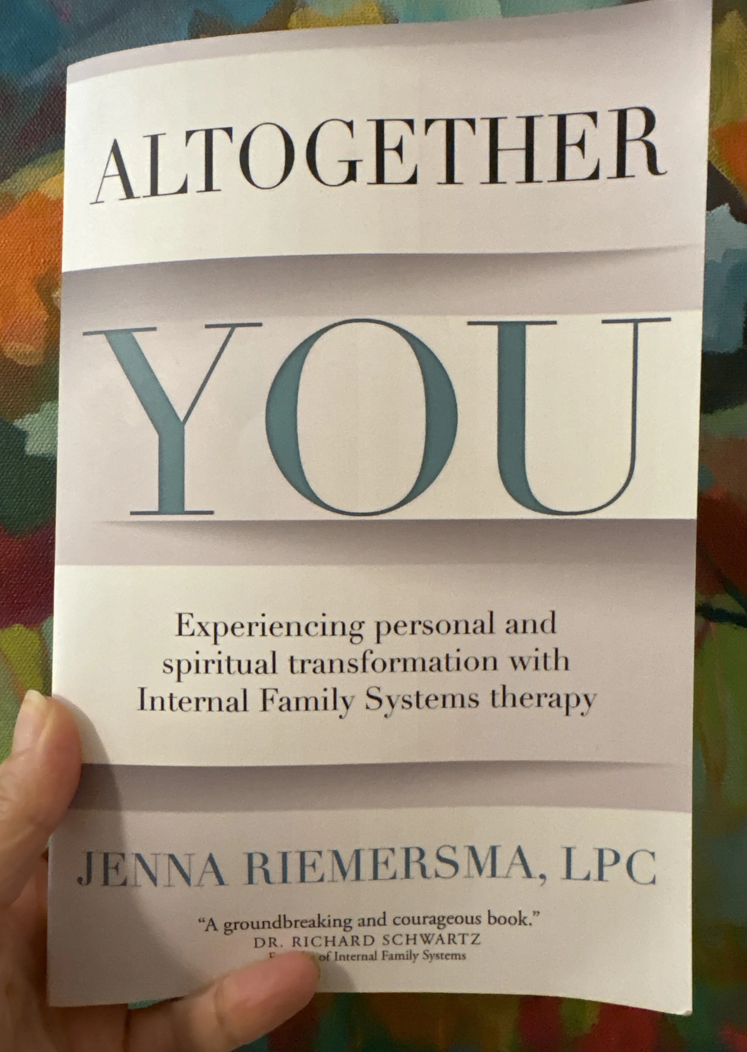 A person is holding a pamphlet that says 'All Together You' at the top, with additional text about experiencing personal and spiritual transformation through Internal Family Systems therapy. The lower part of the pamphlet features a quote praising the book, and the author's name, Jenna Riemersma, LPC. The background includes a colorful, patterned surface.