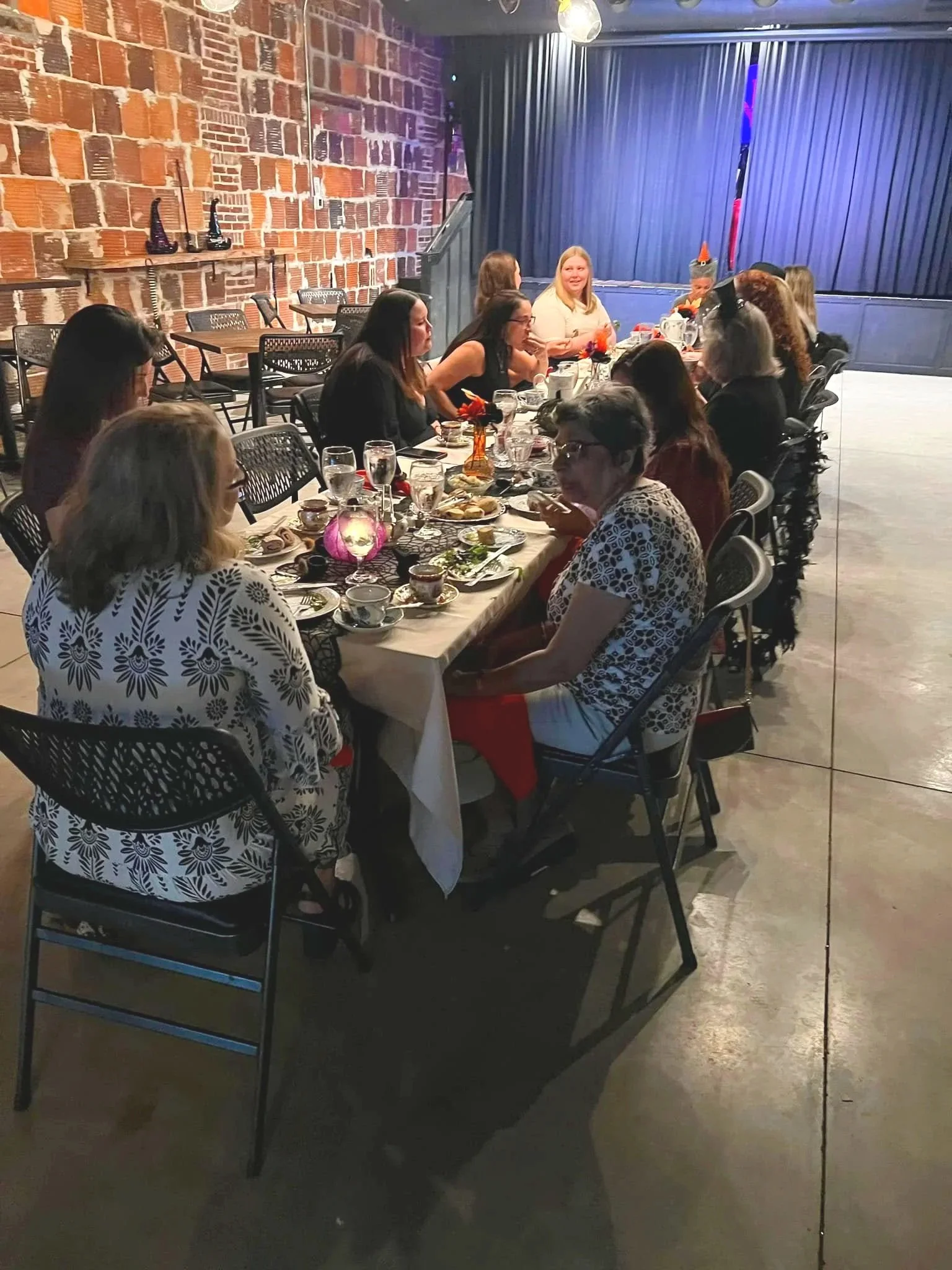 A group of women seated around a long table during a celebration, with some wearing orange party hats, in a brick-walled venue with a stage and blue curtains in the background.