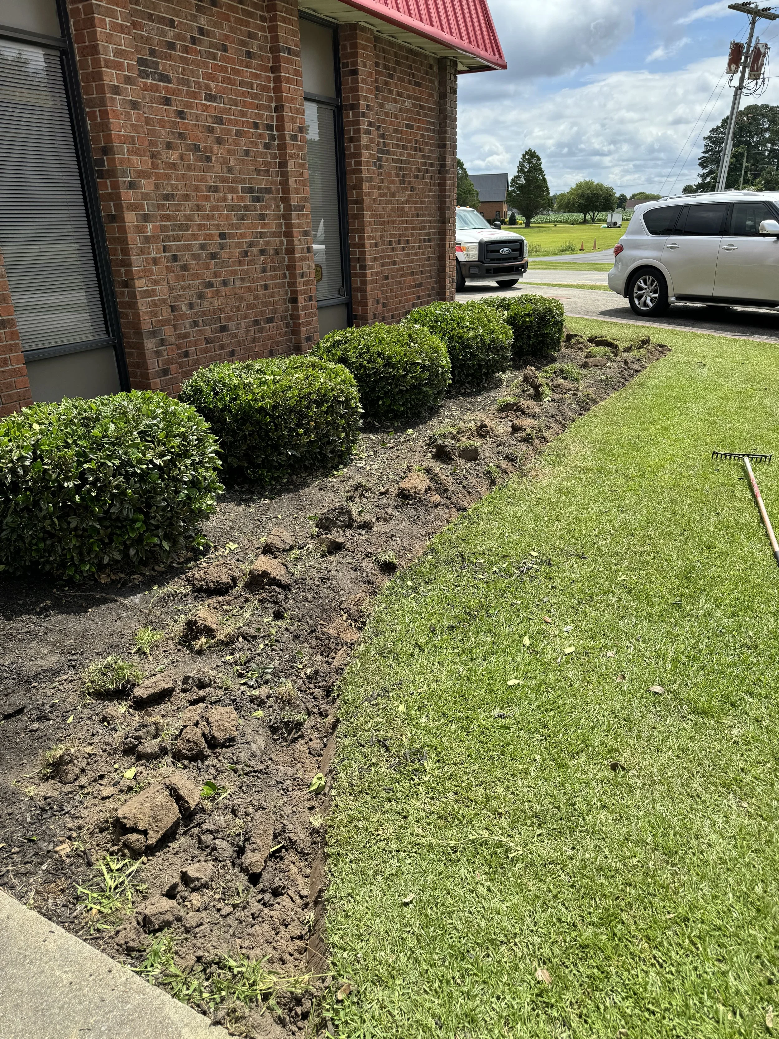 A row of neatly trimmed green bushes planted along a brick building with a newly tilled soil bed in front of them, on a sunny day with partly cloudy skies. A rake is on the grass nearby, and there are two parked cars and some trees in the background.