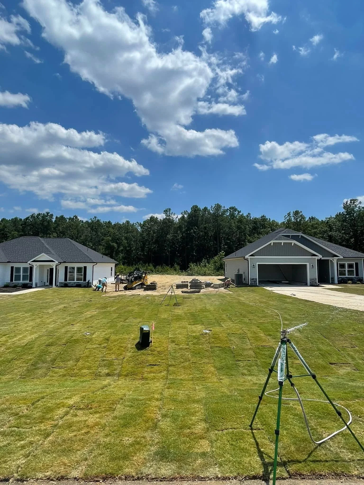 Residential construction site with two houses, workers, and landscaping equipment under a partly cloudy sky.