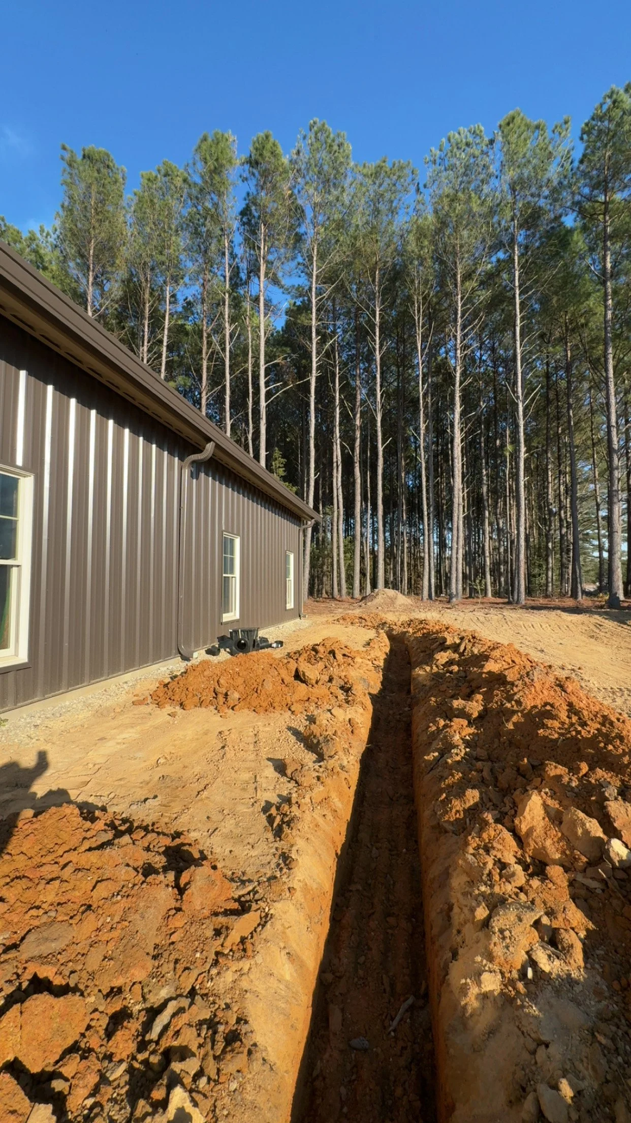 Trench dug beside house with metal siding, in front of a wooded area under a blue sky.