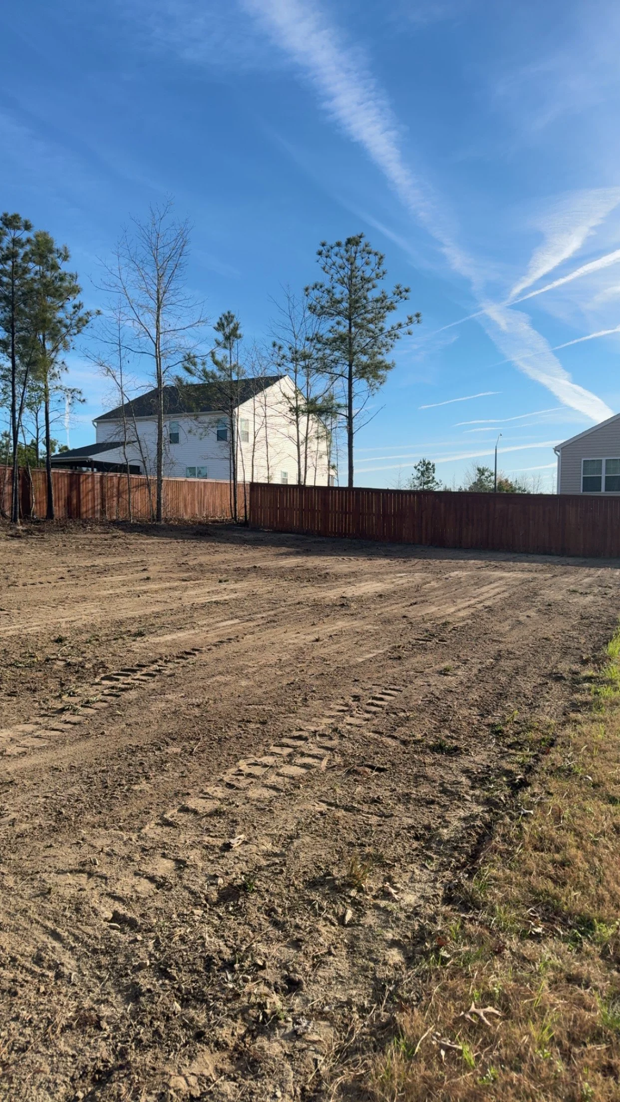 A cleared dirt lot with tire tracks, enclosed by a wooden fence, with trees and a house in the background under a blue sky with streaked clouds.