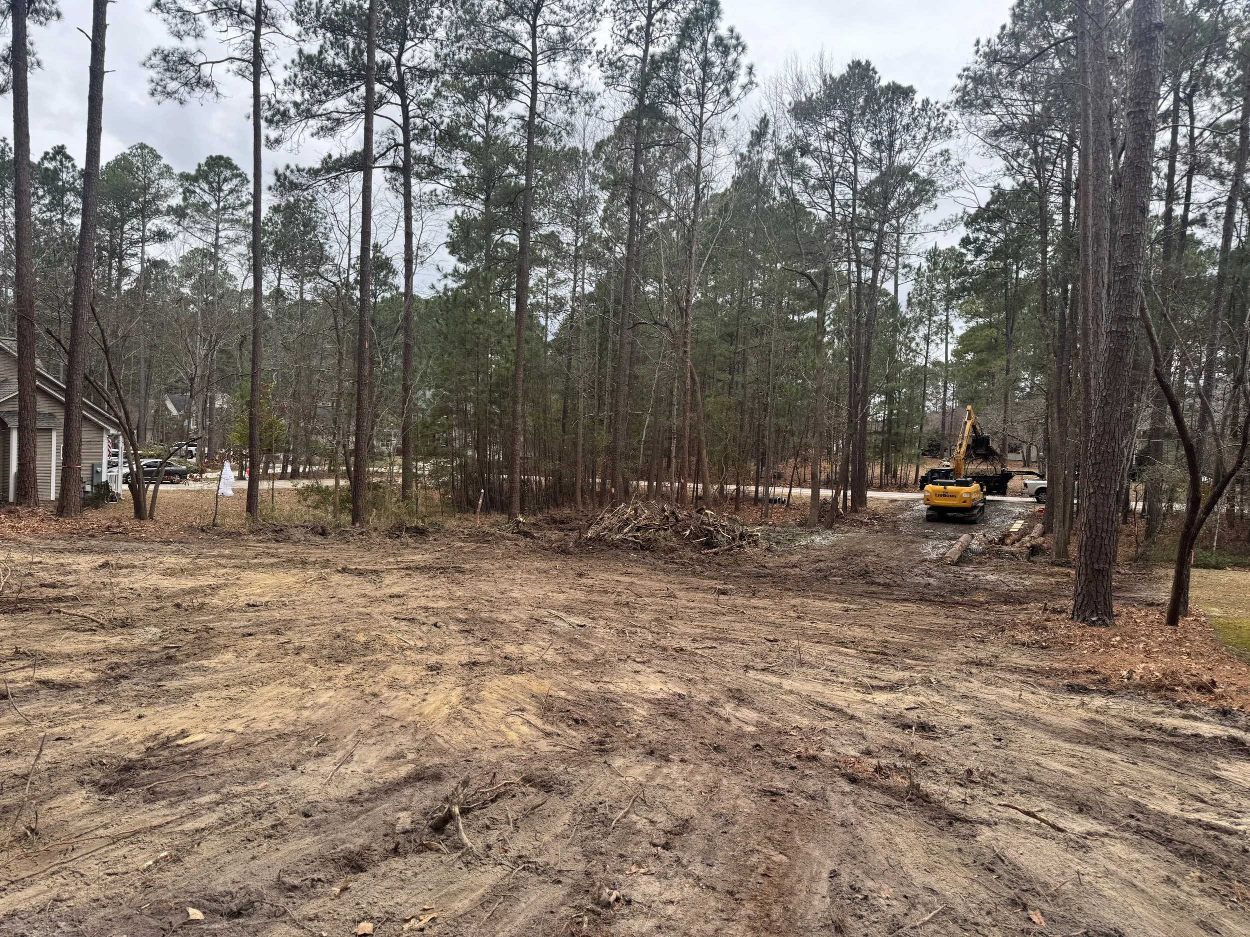 A cleared area of land with dirt ground and tire tracks, surrounded by tall pine trees. In the background, there is construction equipment, including an excavator and a truck, near a residential neighborhood with houses and cars.