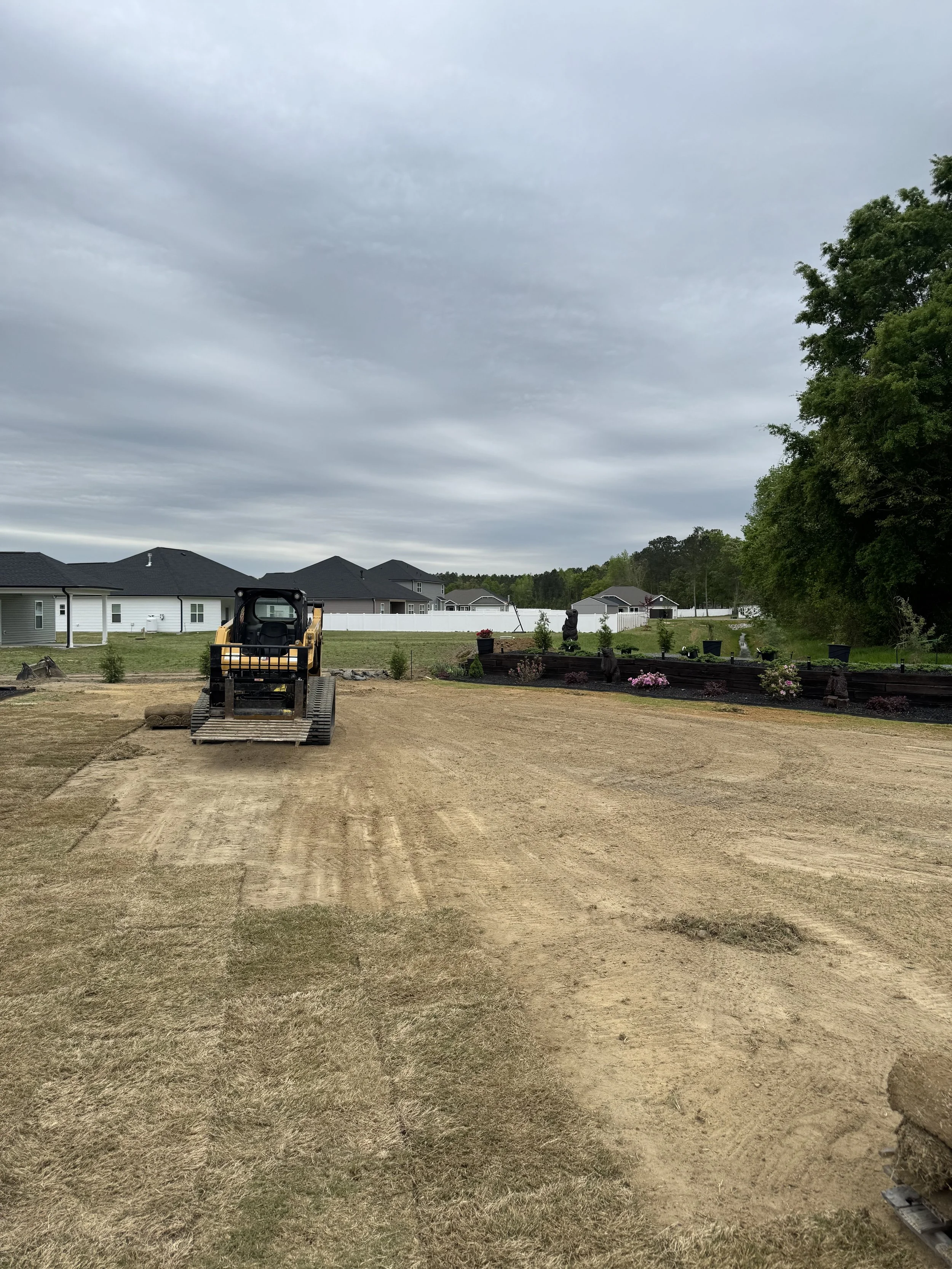 A yellow compact track loader on a dirt pathway in a backyard, with a row of houses and trees under a cloudy sky in the background.
