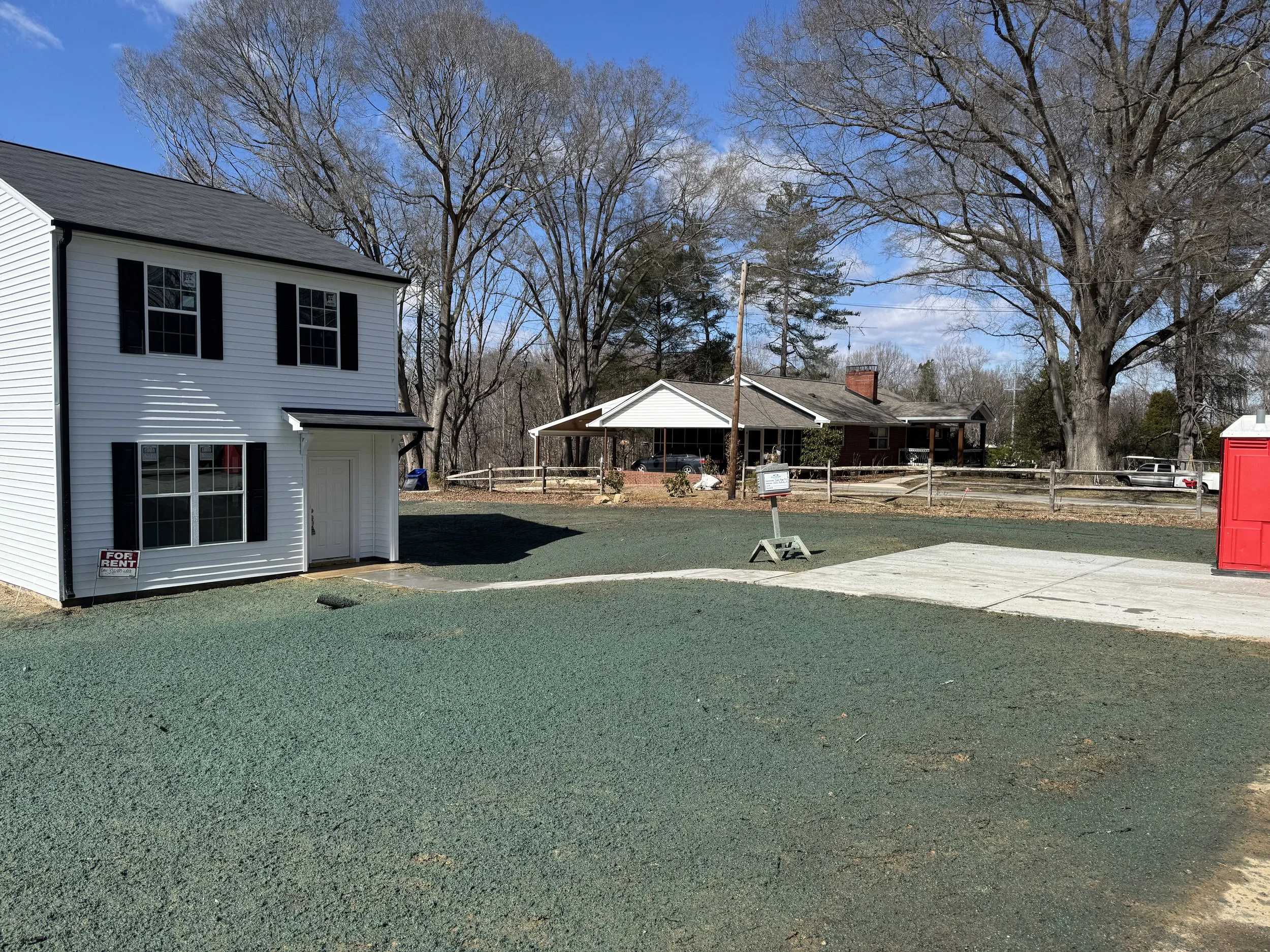 A white two-story house with black shutters and a door on the front. A sign in the yard says for rent. There is a large tree in the background, a concrete driveway, and a red shed on the right.