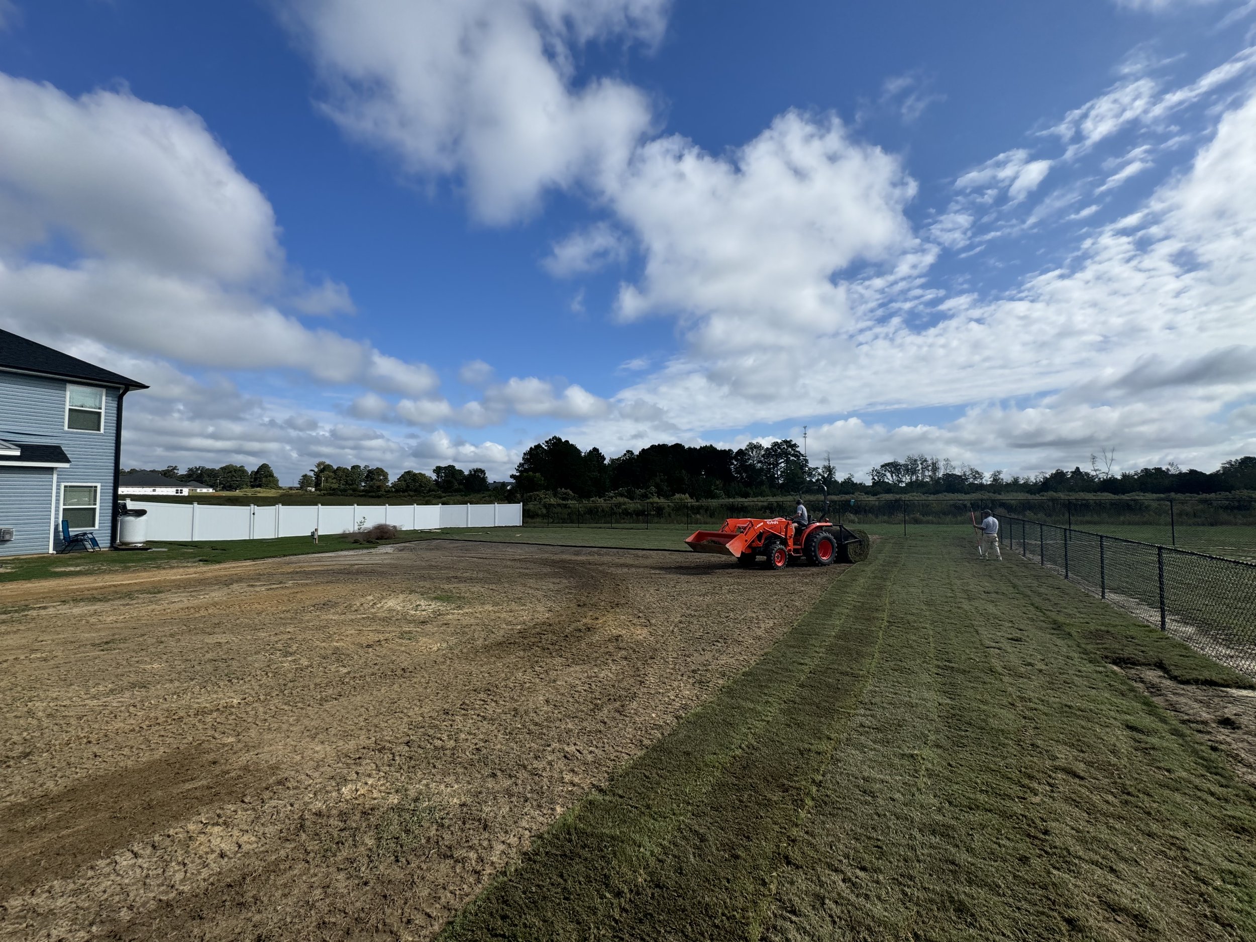 A tractor is leveling soil on a field next to a blue house, with a person walking along the fence on the right under a partly cloudy sky.