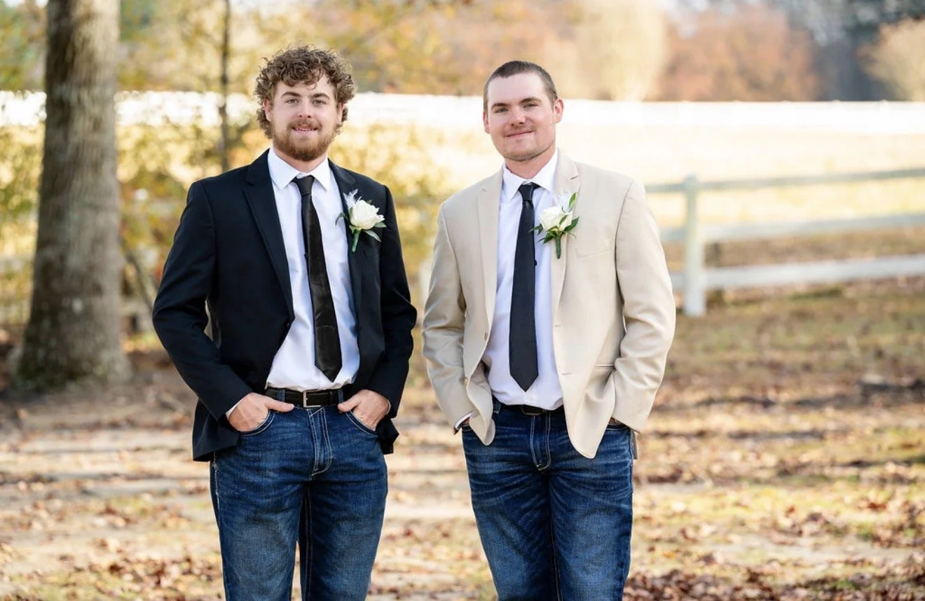 Two men standing outdoors in autumn, dressed in suits with ties and white roses pinned to their lapels, smiling at the camera.