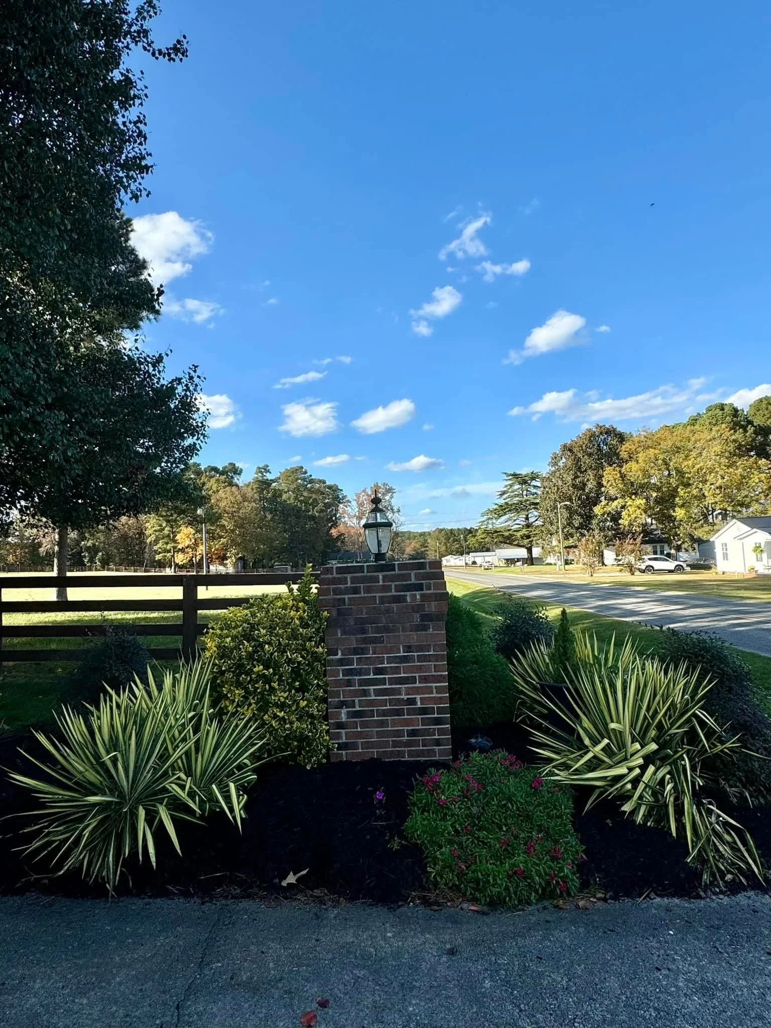 A brick pillar with a lantern on top, surrounded by green bushes and plants, along a sidewalk with a suburban street and trees in the background under a clear blue sky.