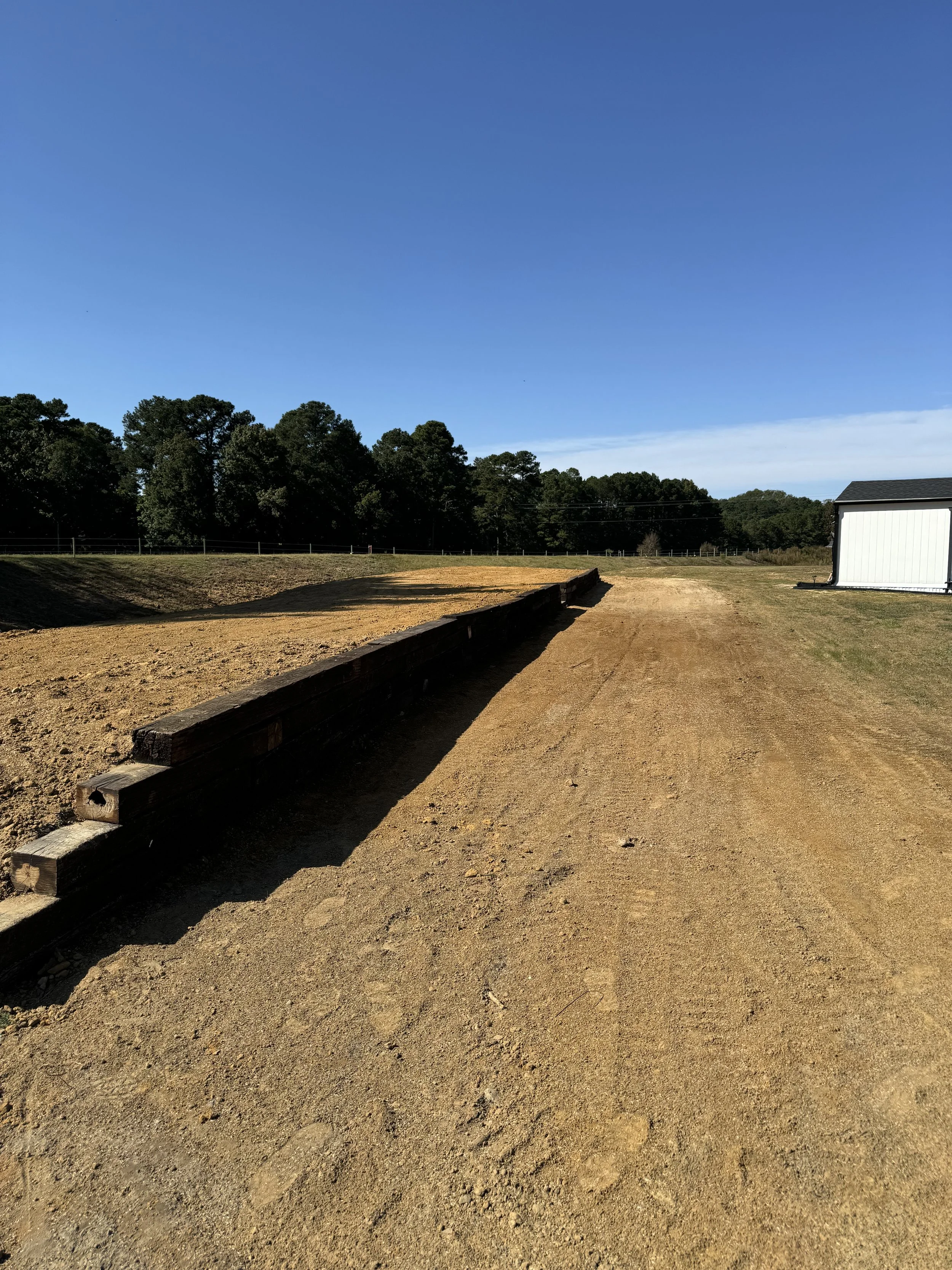 A dirt field with a wooden retaining wall stretching along its edge, a white building to the right, and trees in the background under a clear blue sky.