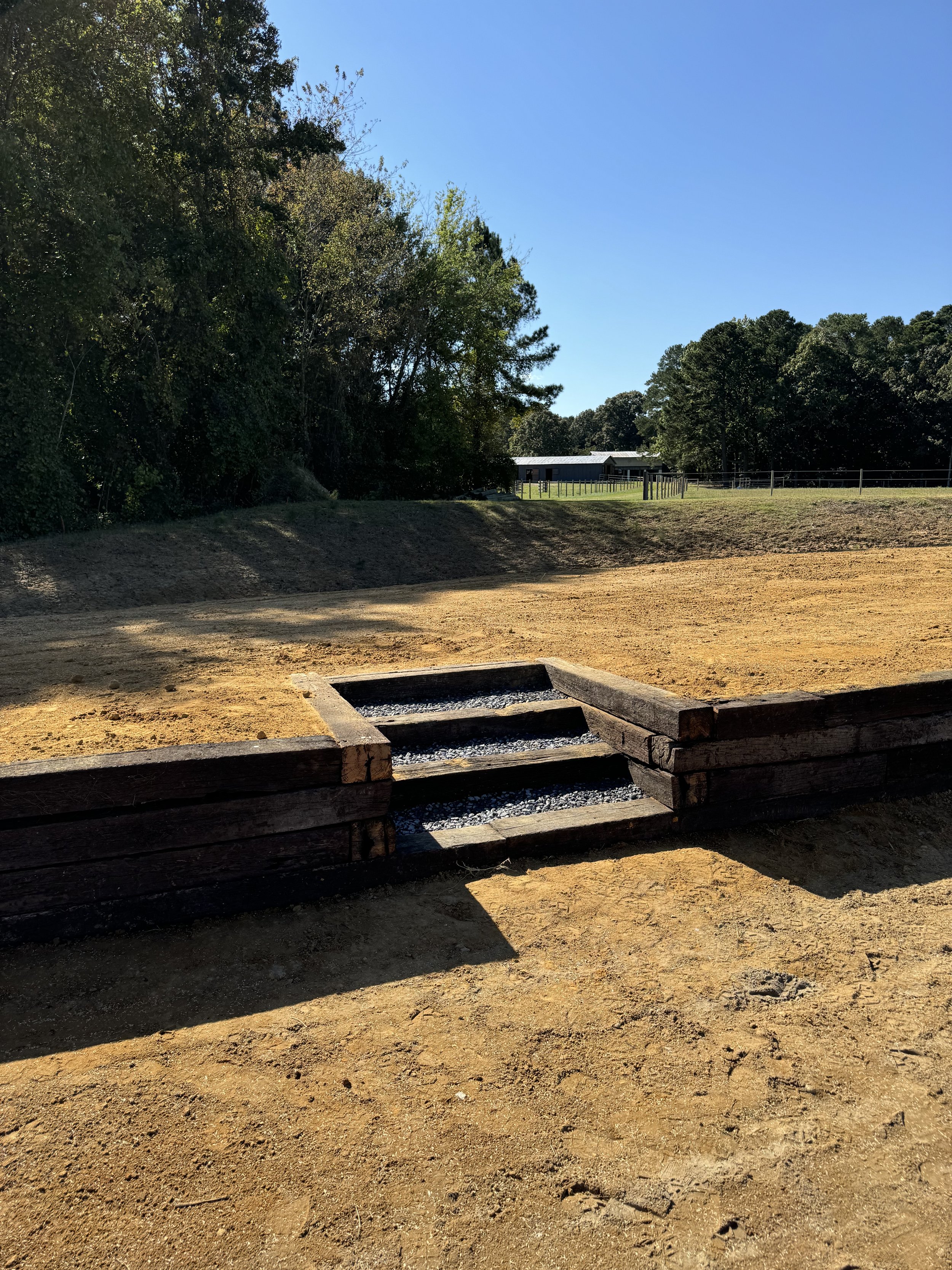A dirt riding arena with a small set of wooden steps leading into a water feature, surrounded by trees and a clear blue sky.